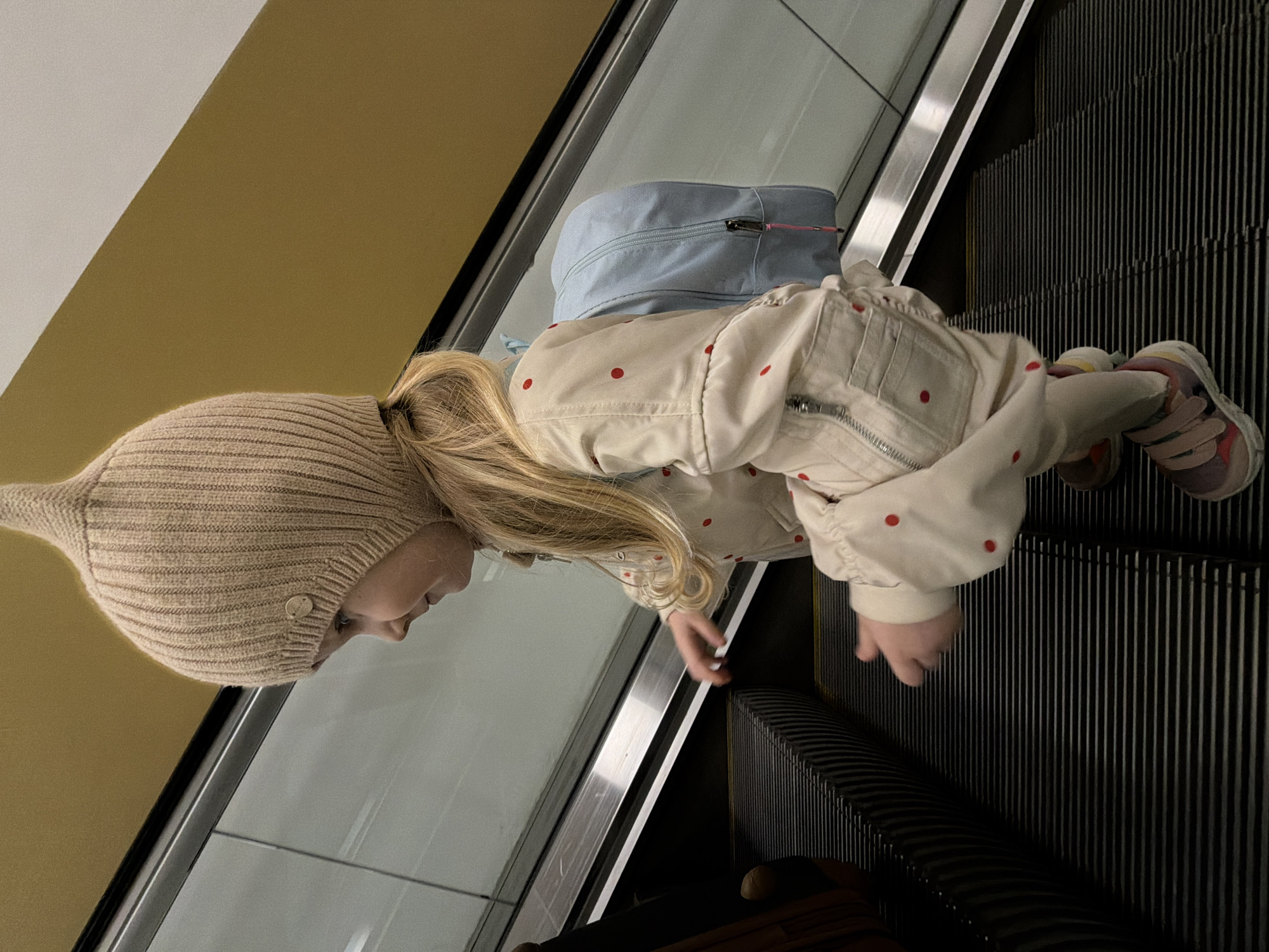 A young child, wearing a tan-colored knit hat with a pointed top, a light-colored jacket with red polka dots, and a light blue backpack, is riding an escalator. The escalator has black steps and a silver railing. The background includes a tan wall and a glass panel. The child is facing upwards on the escalator. There is no evidence that can confirm the location in the city of Khimki, Russia.