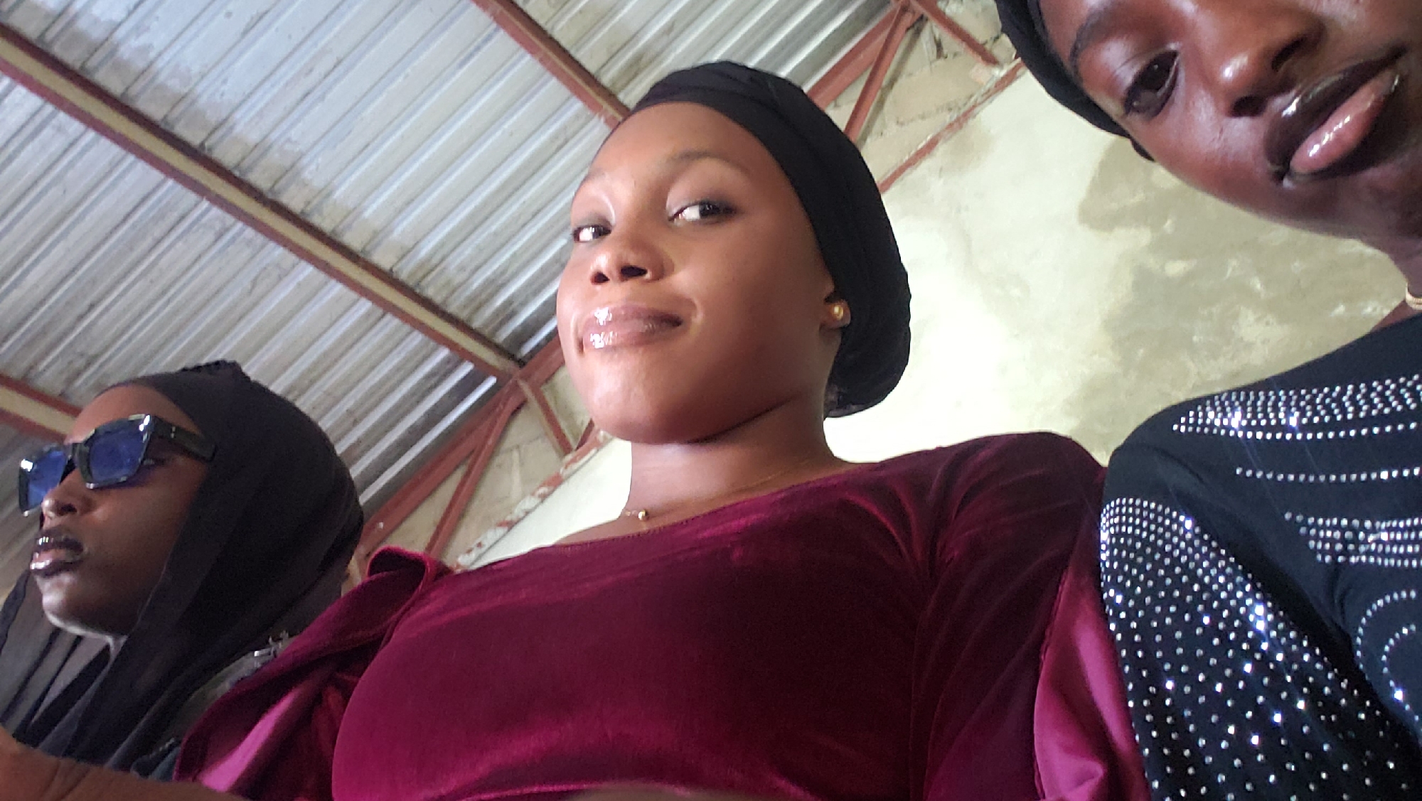A low-angle, close-up photograph captures three young women indoors in Narayi, Nigeria. The central figure, a young woman with medium-dark skin, wears a black head covering and a deep maroon, long-sleeved top, possibly velvet. She has a small golden stud earring on her right ear and a thin gold chain necklace with a small pendant. Her lips are glossy, and she looks directly at the camera with a subtle smile. To her right, partially cropped, is another young woman with dark skin, also wearing a black head covering. Her visible black top is adorned with numerous small, shiny embellishments, forming a patterned design on the shoulder and sleeve. To the left and slightly behind the central figure, a third young woman with dark skin wears a black head covering and large, dark blue rectangular sunglasses, obscuring most of her face. The background reveals an interior space with a corrugated metal ceiling, supported by reddish-brown metal beams and lighter structural elements. The walls are light-colored, appearing to be painted or plastered concrete with some texture. The scene is illuminated by ambient daylight.