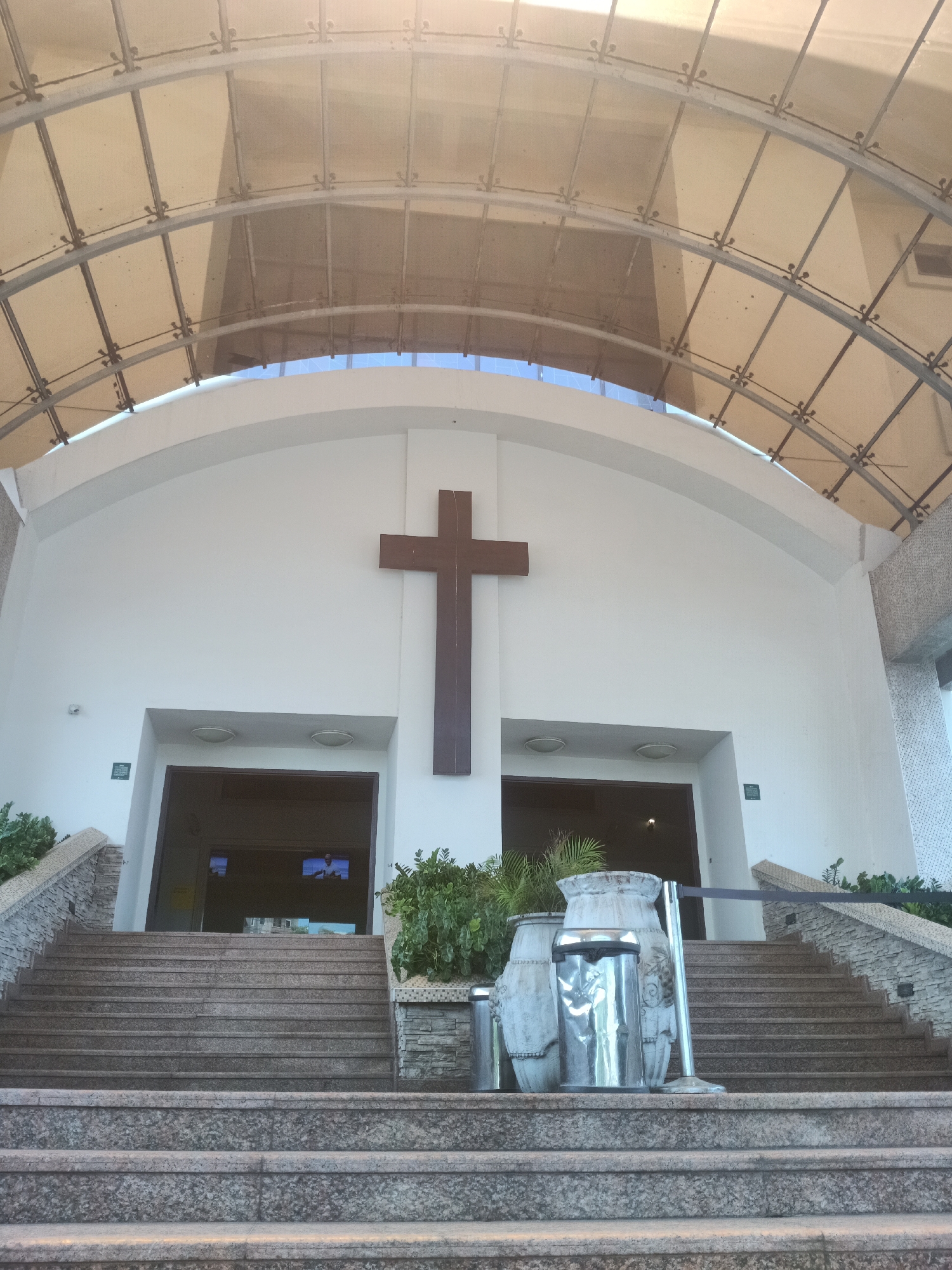 The image shows the exterior of a church in Igboefon, Nigeria. The building is white with a large wooden cross mounted centrally above the entrance. The entrance is accessed via two sets of granite steps leading to separate doorways, with planters flanking each set. A metal railing spans the top of the lower set of stairs. Above the main structure, a curved, translucent canopy with a metal frame covers the entrance area.