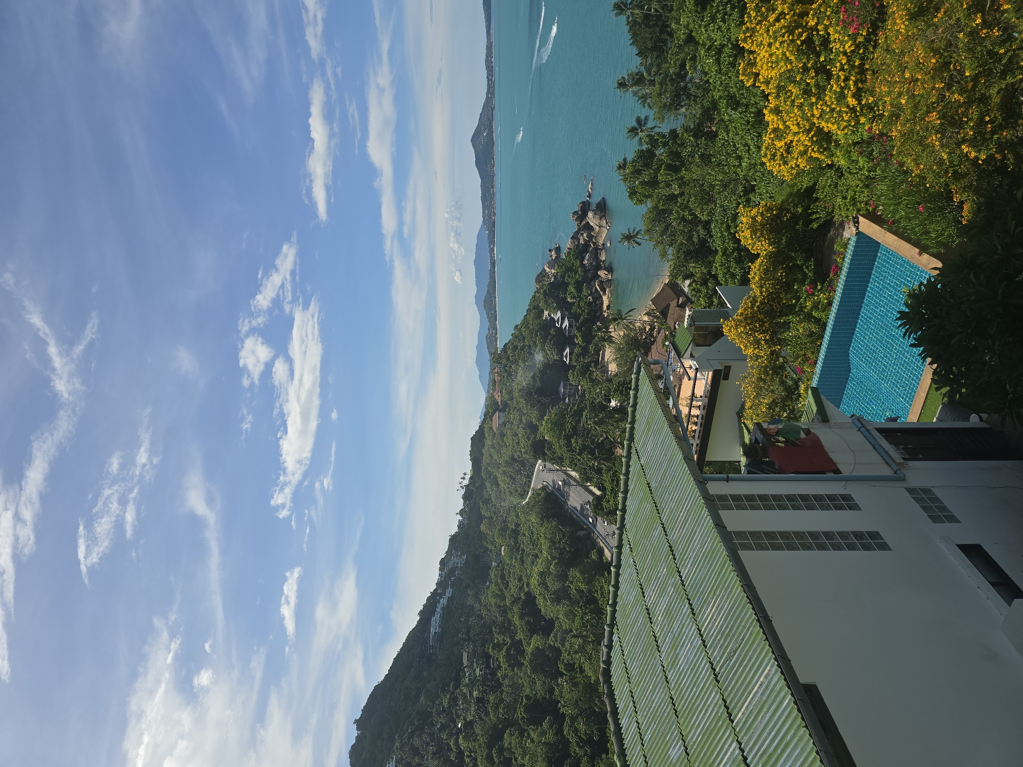 The image captures a vibrant coastal landscape under a bright, cloudy sky. In the foreground, a white building with a green corrugated metal roof is visible, featuring glass block windows and a small balcony. Adjacent to the building is a bright blue swimming pool surrounded by lush green foliage and a burst of yellow flowering bushes.
The middle ground reveals a winding road cutting through a densely forested green hillside, dotted with buildings. The road is traversed by several vehicles. Further down, the hillside slopes towards the turquoise ocean. A small beach with scattered rocks and palm trees lines the shore, leading to the expanse of the sea.
In the distance, the ocean stretches out, with several boats leaving white wakes on the water. The horizon is marked by a series of mountains and islands under a sky filled with scattered white clouds against a backdrop of clear blue. The overall impression is a tropical paradise with a mix of man-made structures and abundant natural beauty.