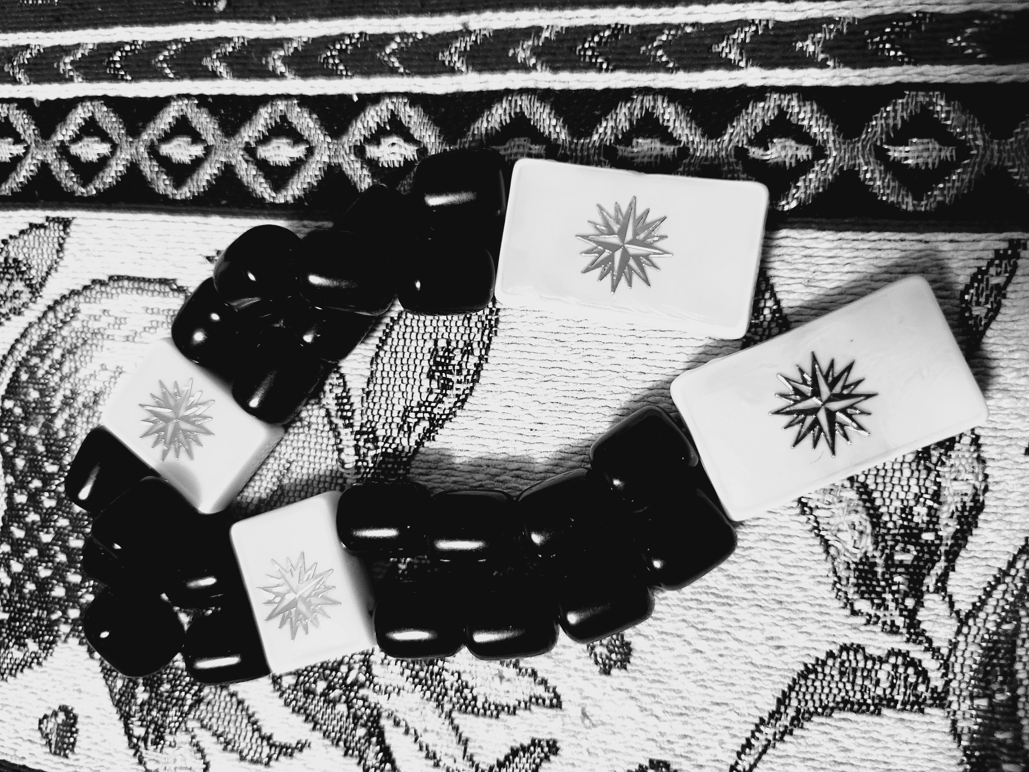 The image shows a collection of objects on a textured surface. There are four rectangular white tiles, each decorated with a starburst-like symbol, interspersed with a collection of small, dark, polished stones. The background appears to be a fabric with a complex woven pattern, likely a tablecloth or rug. The style of the pattern suggests it could be a traditional Armenian design. No specific landmarks are visible to confirm the location as Yerevan, Armenia; the statement is based on the style and potential cultural context of the scene.