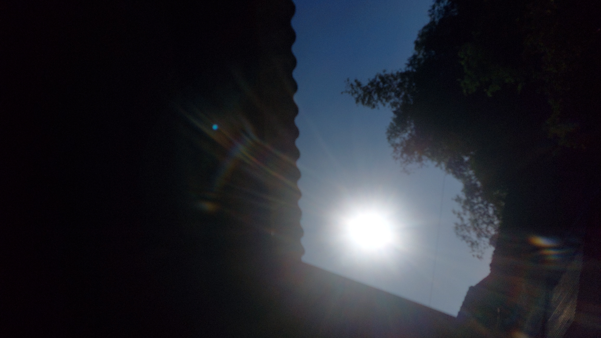 The image depicts a bright sun shining in a clear blue sky, partially obscured by a dark, corrugated roofline in the foreground. Sunlight causes lens flare, creating streaks of light and rainbow effects. Silhouetted trees and a building are visible against the sky and in the darker foreground. The overall scene suggests an outdoor view from a shaded area, possibly a home in Jatirogo, Indonesia, during daylight.