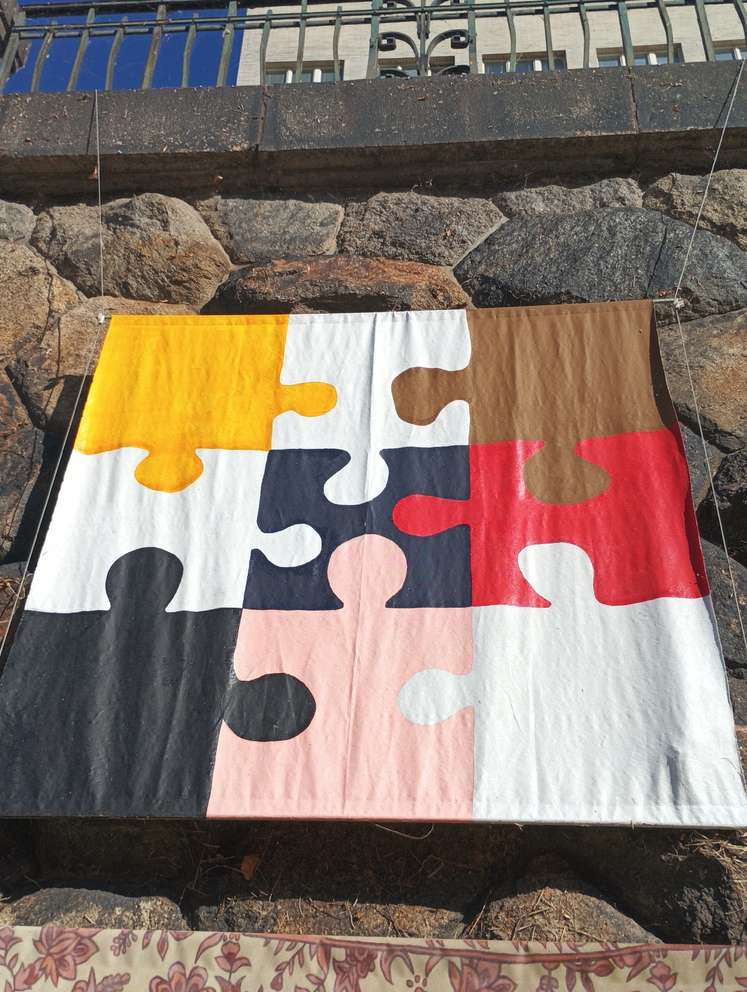 An outdoor scene in Pilsen, Czechia features a large, canvas-like object resembling a jigsaw puzzle, suspended against a stone wall. The puzzle pieces are painted in various solid colors: yellow, brown, red, black, pink, and white. The puzzle is hung using wires, with one wire visible extending from the top left corner of the puzzle and another from the top right. Behind the stone wall, a dark metal railing with decorative elements is visible, topped by a building with windows. The foreground shows dry grass and dirt.