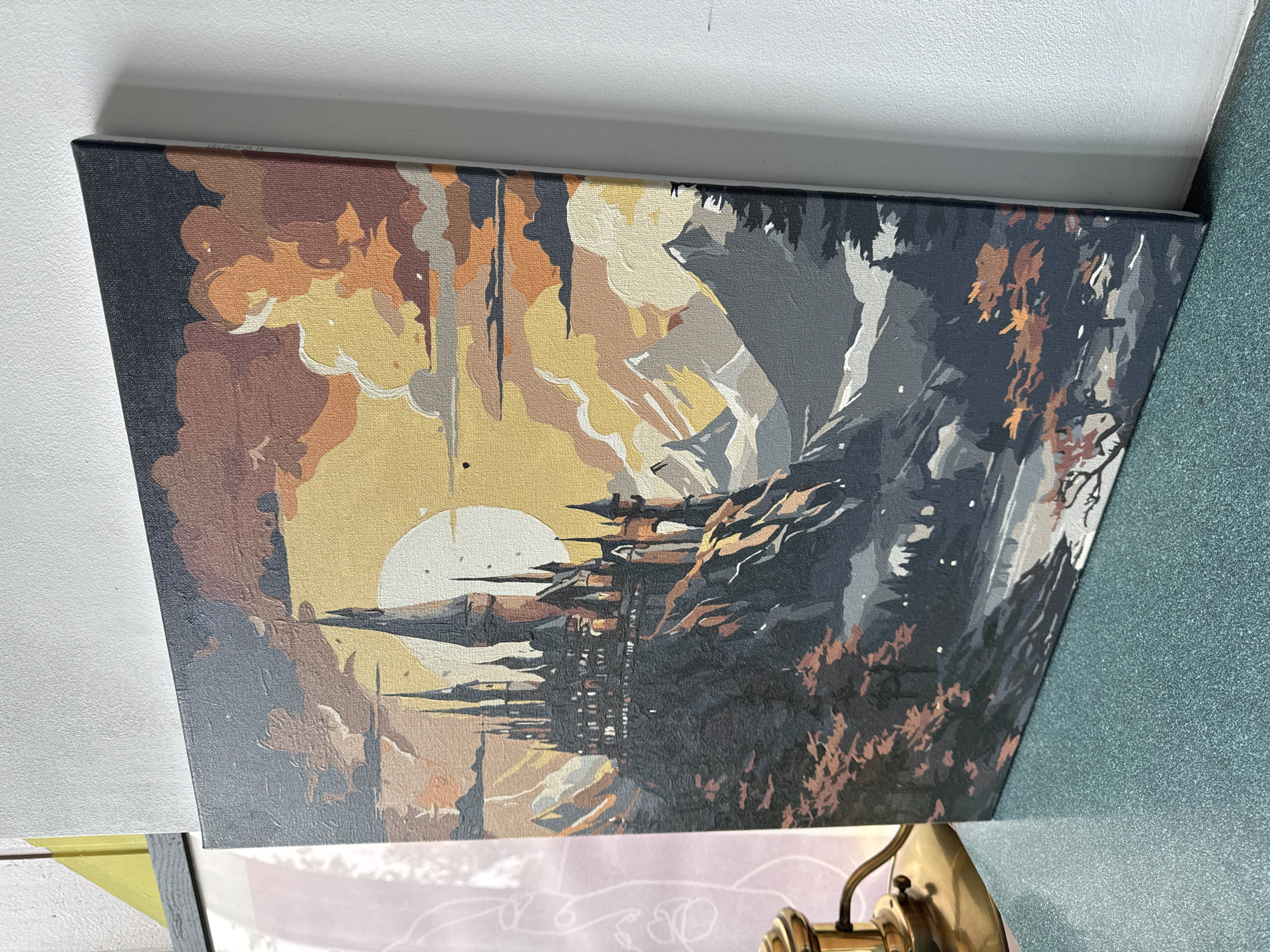 The image depicts a paint-by-numbers canvas print of a landscape. The subject is a fantastical city or castle perched on a rock formation. The overall color palette is muted, with shades of brown, yellow, and gray. A stylized sun is visible in the background. The artwork has a textured appearance due to the canvas and paint application. The canvas is leaning against a wall.