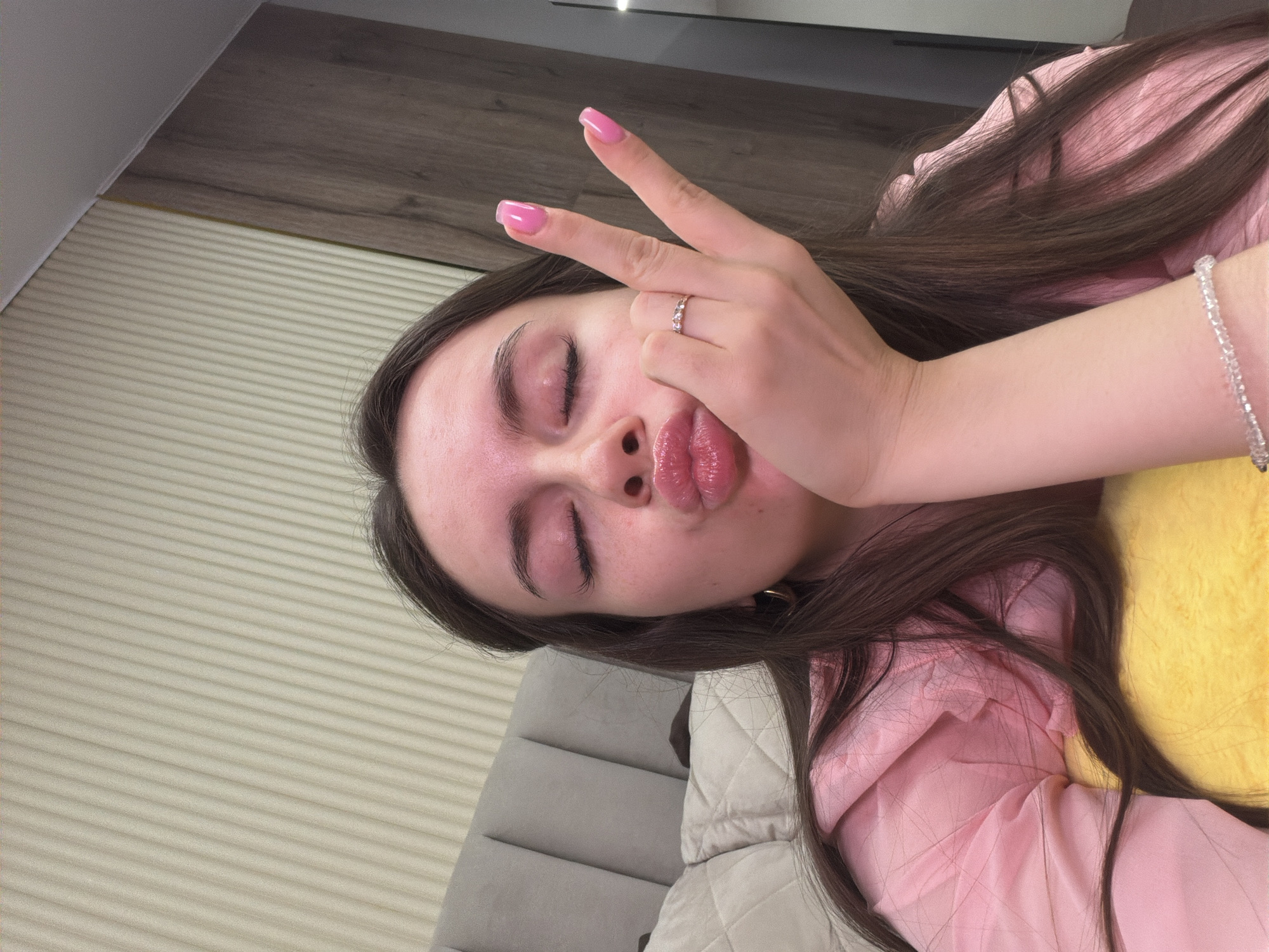 The image displays a young adult female captured in a close-up, selfie-style indoor shot. She has long, dark brown hair, closed eyes, and her lips are pursed in a glossy pink "kissing" gesture. Her left hand (viewer's right) is raised, forming a "peace" sign with two extended fingers, showcasing bright pink manicured nails and a ring with several small stones on her ring finger. Her right hand (viewer's left) supports her chin. She wears a pink top and a clear, beaded bracelet on her right wrist. A soft, yellow object is partially visible beneath her. The background behind the subject includes a light gray upholstered headboard of a bed, a cream-colored wall with vertical ribbed paneling, and a section of dark brown wooden paneling, indicating an interior residential setting. The location for this scene is Chaikovsky, Russia.
