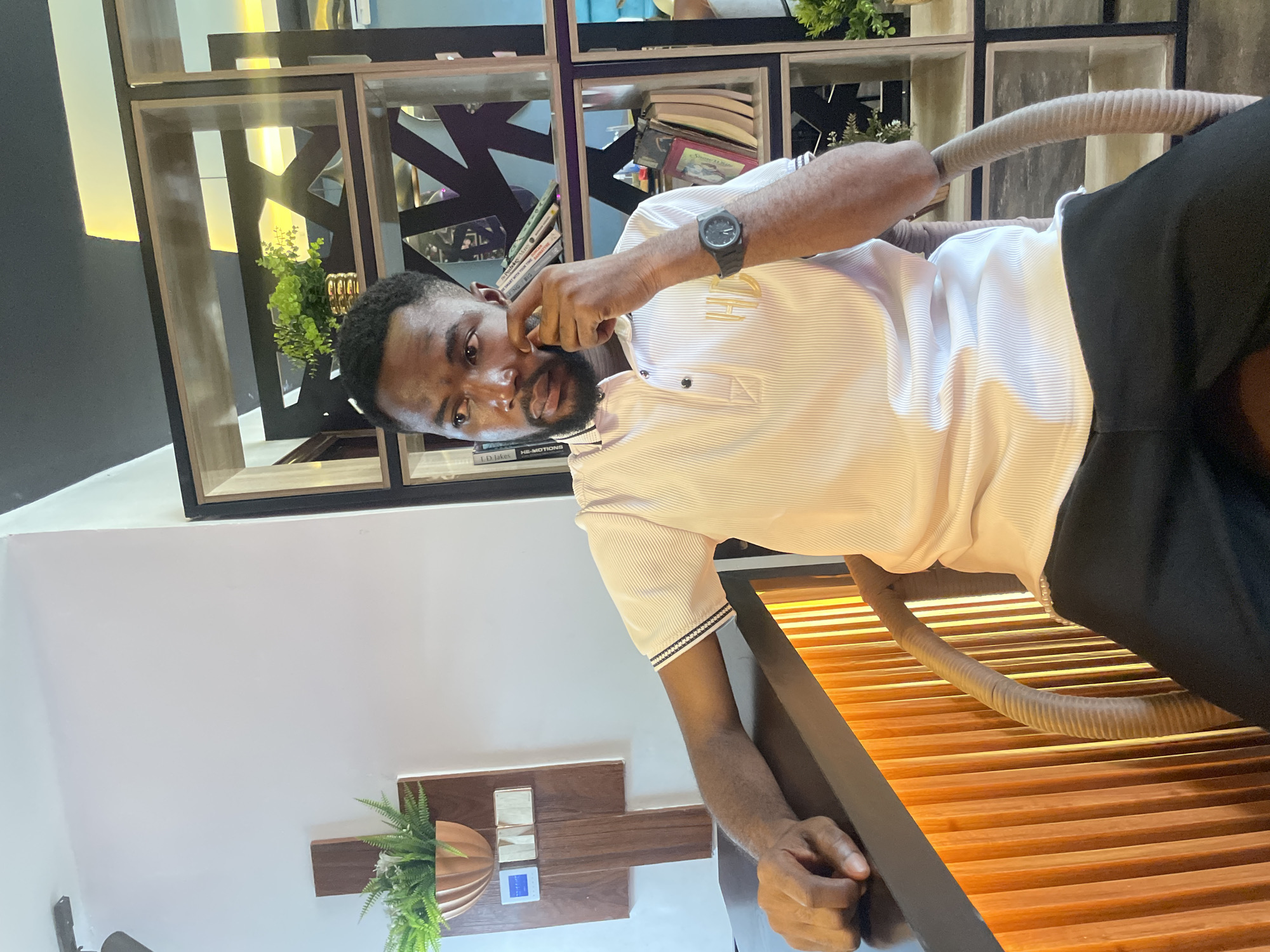 A man is sitting in a chair indoors. He is wearing a white striped short-sleeved shirt with gold lettering on the chest, a black watch on his left wrist, and black pants. He is touching his face with his right hand.
Behind him is a shelving unit with various objects, including books and plants. The wall is white and has a wooden decorative piece with a plant. The chair appears to be wooden with a tan cushion.
The image may have been taken in Adankolo, Nigeria, based on the user's prompt, but there is no discernible information in the image to confirm this location.