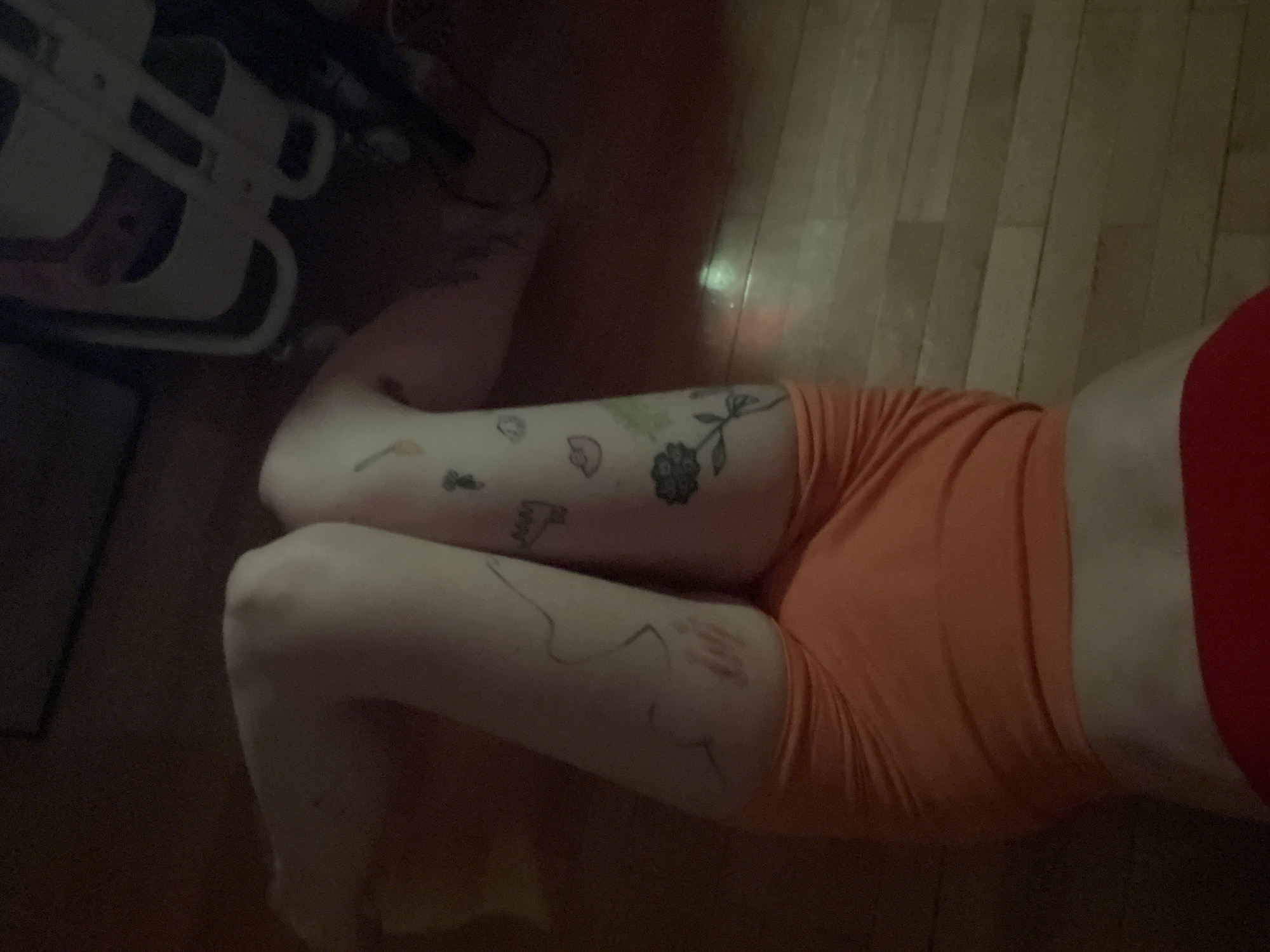 The image provides an overhead view of a person lying on a light-colored wooden floor. The visible parts of the person include their upper torso, clad in a red top or bra, and their legs, dressed in orange shorts. Their knees are bent, and the legs are positioned together.
The person's legs are adorned with multiple tattoos. On what appears to be their left thigh (on the right side of the image), there is a detailed floral tattoo, resembling a rose with leaves, alongside several smaller outline tattoos depicting a mushroom, a heart shape, an arrow, and other subtle designs. On their right thigh (on the left side of the image), intricate line drawings are visible, appearing as abstract figures or shapes, possibly a stylized face or silhouette, and a design that resembles a handprint or gestural drawing.
The setting is indoors, featuring a wooden floor with visible planks. The lighting is dim and soft, casting some reflections on the floor, suggesting a low-light environment, possibly evening. In the top-left corner, a dark and blurry fragment of a white, possibly tubular or framed, object or piece of furniture is present. The overall scene conveys a moment of rest or relaxation. No discernible text is visible on the person or in the immediate surroundings. The location context for this image is Belgrade, Serbia.