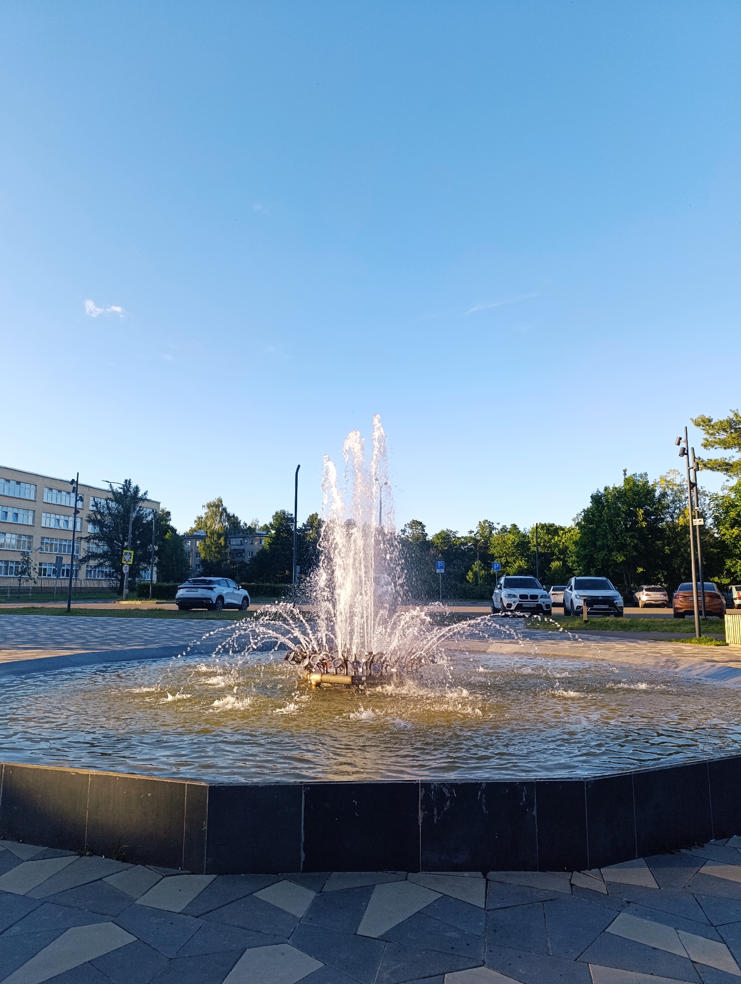 The image depicts a tranquil outdoor scene in Chernogolovka, Russia, featuring a large, active fountain at its center. The fountain sprays water upwards and outwards, creating a dynamic focal point within a circular pool. The pool has a dark, stone-like rim and is surrounded by a paved area with an interesting geometric pattern.
In the background, a multi-story building with many windows and a light-colored facade is visible, suggesting an urban or suburban environment. Behind the building and to the right, a line of lush green trees adds a natural element to the scene. Several cars are parked on the side of a road in front of the trees, indicating the presence of a public space or parking lot.
The sky is a clear, vibrant blue with a few wispy clouds, and the sunlight suggests it is either morning or late afternoon. The overall atmosphere is peaceful and serene, with the gentle sound of the fountain likely contributing to the calmness. There are no people visible in the frame. The location context indicates this is Chernogolovka, Russia.