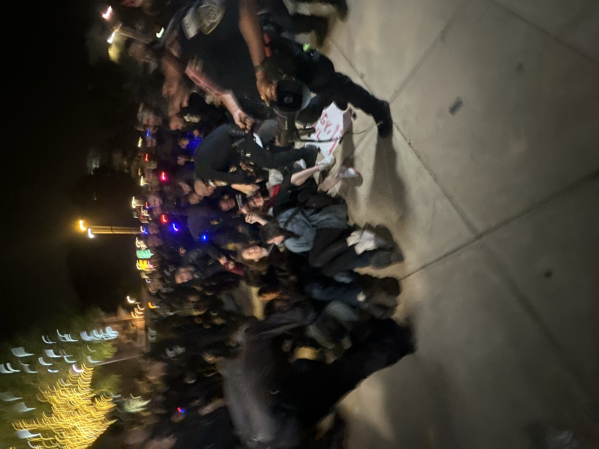 The image shows a large group of people gathered at night. Some individuals appear to be lying on the ground, while others are standing around them. Several figures are holding megaphones. The location is not specified in the image.