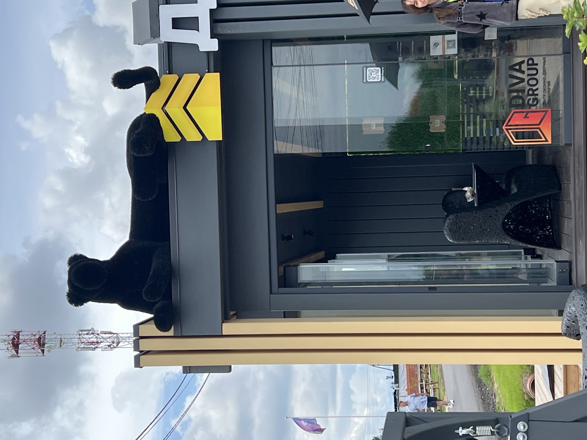 In Yakunino, Russia, a black plush panther sculpture is perched atop a modern building, its body stretched horizontally. Below the panther, a large yellow chevron-like symbol is visible. The building itself is dark grey with vertical siding and features large glass doors. A sign on the glass reads "DIVA GROUP" with smaller text below in Russian. To the right, a young woman wearing a grey jacket and light-colored pants stands with an umbrella. In the background, a telecommunications tower with red and white accents pierces the cloudy sky. A purple and red flag flies on a pole to the left. A man in a light blue shirt and dark shorts walks on a paved path in the distance, near some outdoor seating. The environment suggests an outdoor commercial or exhibition space.