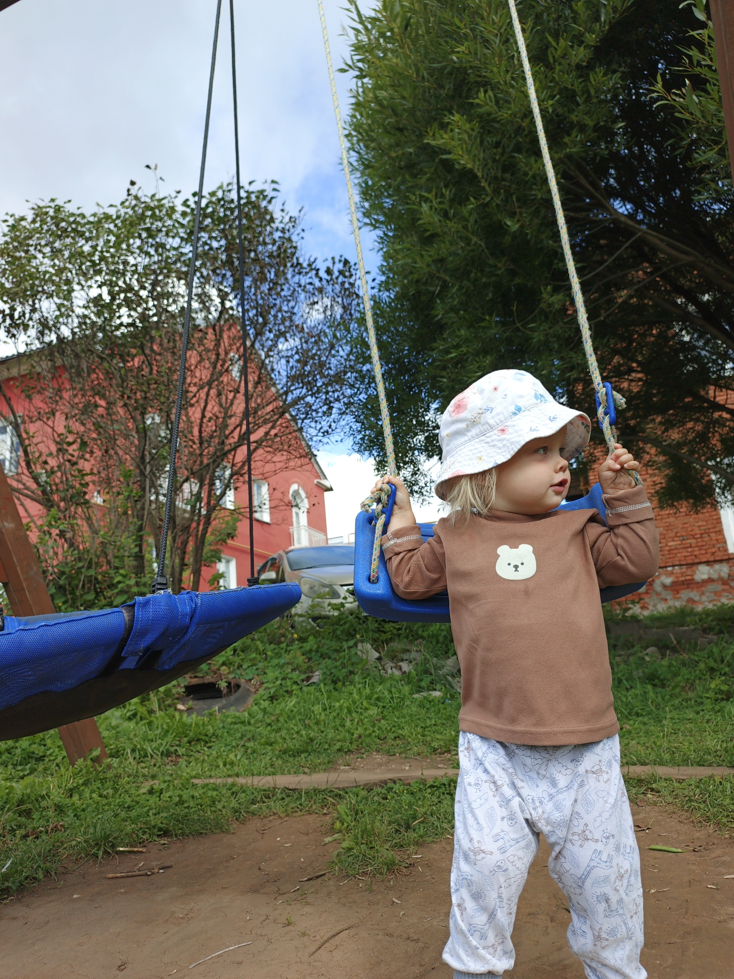 A young child wearing a white bucket hat with a floral pattern and a brown long-sleeved shirt with a bear appliqué stands on a dirt path in front of a red brick building in Medvedyovka, Russia. The child is holding onto the ropes of a blue swing set. The background shows trees, grass, and the red building, which appears to be a house with white trim and windows. The sky is overcast with patches of blue. The scene depicts an outdoor setting, likely a backyard or playground, on a cloudy day.