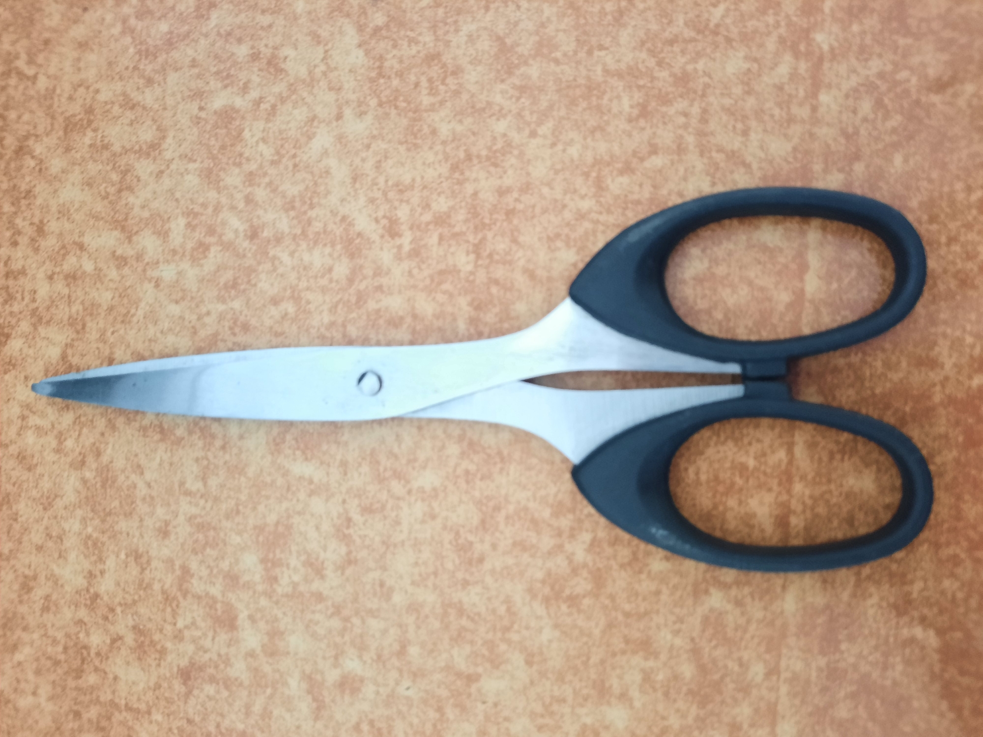 A pair of open, silver scissors with black plastic handles lies on a textured, light brown surface. The scissors are positioned vertically, with the blades pointing upwards and the handles downwards. The blades are sharp and pointed, and they are slightly angled away from each other. The handles are oval-shaped and appear to be made of a smooth, matte material. The background surface has a mottled pattern of lighter and darker brown tones, resembling wood grain or a textured countertop. There is no indication of the city of Lhokseumawe, Indonesia, in the image.