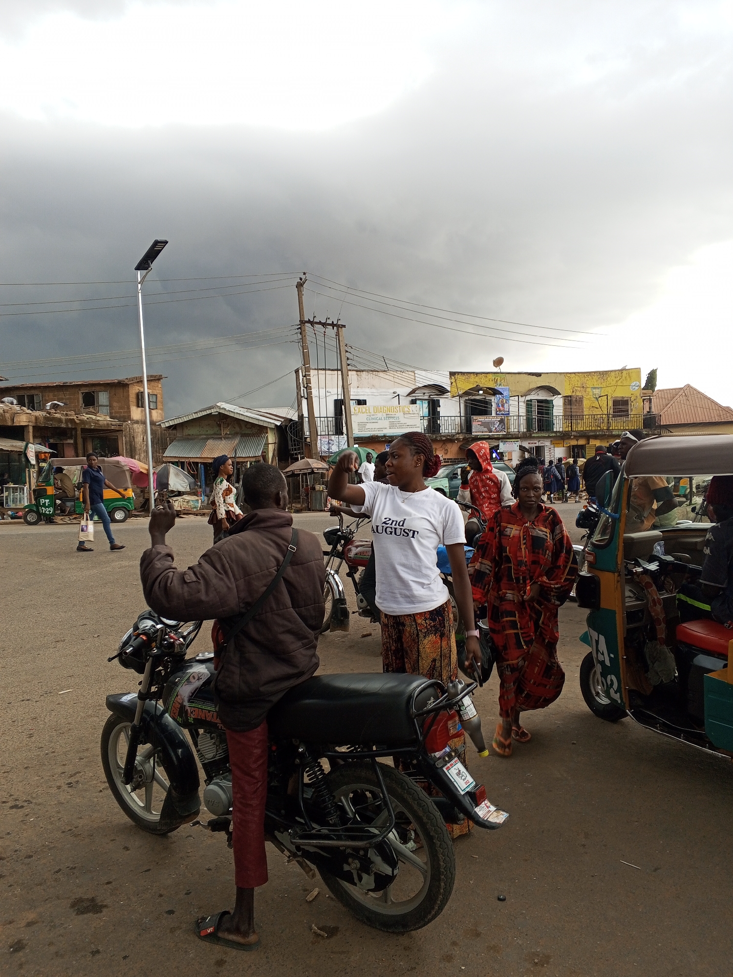 The image depicts a street scene in Gwong, Nigeria, under a cloudy sky. In the foreground, a man sits on a black motorcycle, facing away from the camera. He is wearing a brown jacket and red trousers. Next to him, a woman in a white t-shirt with the text "2nd August" and a patterned skirt is gesturing with her right hand. Another woman in a red patterned dress walks behind her. A green and red tricycle (auto-rickshaw) is partially visible on the right side of the frame, with passengers inside. In the background, there are various buildings, utility poles, and pedestrian traffic, typical of a bustling urban area. The overall environment suggests an overcast and possibly humid day.