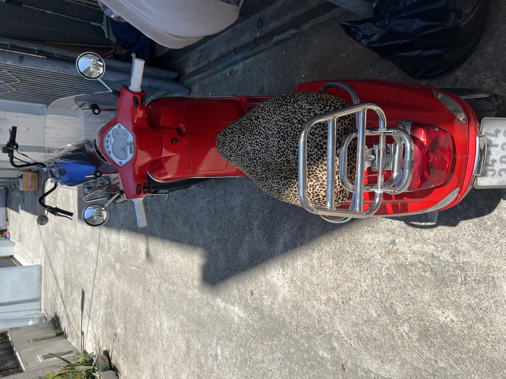 The scene depicts a narrow, concrete alleyway in Nha Trang, Vietnam, illuminated by bright sunlight, casting distinct shadows.
In the foreground, a red scooter, styled similarly to a Vespa, is parked facing away from the viewer. Its seat is covered with a leopard-print fabric, and a chrome luggage rack is mounted over the rear fender. The rear license plate, partially visible, displays "34-TL 2023". The scooter's front features white handlebar grips, a white-faced speedometer, a clear windscreen, and chrome rearview mirrors reflecting portions of the alley.
Directly behind the red scooter, a blue motorcycle, characterized by raised handlebars and a visible fuel tank, is parked.
To the left, a light-colored building wall with small, recessed rectangular elements lines the alley. A potted plant in a decorative ceramic pot rests at the wall's base. To the right, a dark, corrugated metal gate or door is visible against another building. Adjacent to the gate, a blue broom leans against the wall, and a dark blue tarp or fabric is present on the ground. A black plastic trash bag rests on the concrete near the rear of the red scooter. No people or animals are visible, and no actions are taking place.
