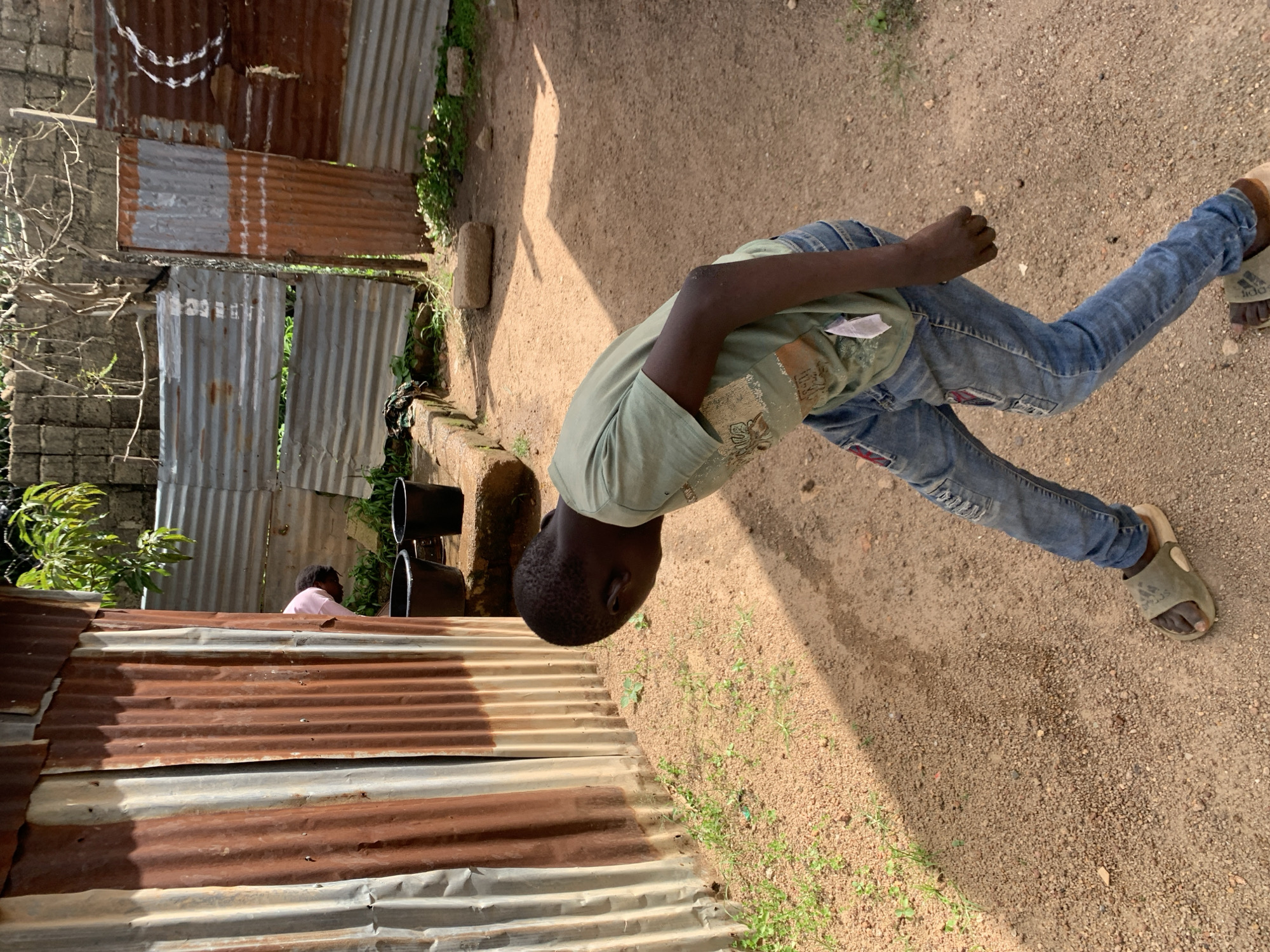 The image shows a young boy wearing a green t-shirt and blue jeans, standing in an outdoor setting. He is bent over, looking down at the ground, with his right arm extended and hand clenched. He is wearing sandals. To the left of the boy is a wall made of corrugated metal sheets, some of which appear rusted. Behind the corrugated wall, a smaller figure, possibly another child, is visible in a pink shirt. The ground is sandy with patches of dry grass. In the background, more corrugated metal structures are visible. The lighting suggests it is daytime, with shadows cast on the ground, indicating sunlight. The overall scene appears to be in a rural or informal settlement, consistent with a location context of Jos, Nigeria. There is no visible text in the image.