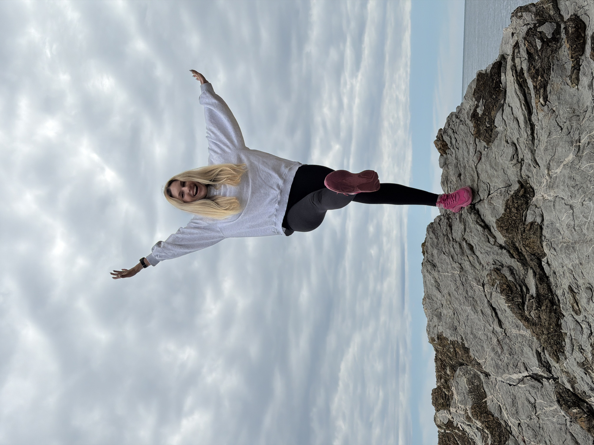 A photograph displays a blonde woman balancing on the edge of a rocky cliff in Budva, Montenegro. She is positioned sideways in the frame, with the cliff edge to the right and the cloudy sky to the left. The woman has one foot securely planted on the rock, and her body is aligned horizontally, mimicking the sky and the sea line in the background. Her arms are extended outwards, as if she is in a flying pose. She is wearing a light grey sweater, dark leggings, and bright pink sneakers. The rock face is detailed with various shades of grey and dark textures, showing rugged terrain. Behind the cliff, a sliver of ocean meets the horizon, and the sky above is overcast with grey clouds.