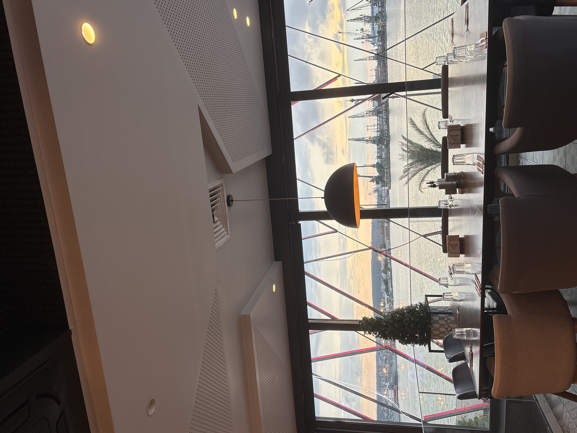 The image depicts the interior of a modern restaurant or cafe with a panoramic view of a large body of water and an industrial port. The perspective is from inside the establishment, looking out through expansive windows.
The interior features a contemporary design with clean, light-colored walls, some of which are textured with perforated acoustic panels. Recessed circular lights emit a warm glow from the ceiling and walls. A unique, dark, semi-circular pendant lamp with an orange-gold interior hangs centrally over a long, dark dining table. The table is set with multiple upholstered chairs in varying shades of brown and gray, clear drinking glasses, and cutlery. A small potted plant in a decorative blue and white pot serves as a centerpiece, alongside condiment shakers. An air vent is visible on a white wall.
Through the large windows, a stunning outdoor scene unfolds during what appears to be either sunrise or sunset, characterized by soft, warm light painting the sky in hues of orange, yellow, and muted blue. The view overlooks a vast expanse of water, likely a sea or ocean, with a distant coastline. A prominent feature of the vista is an active industrial port, teeming with numerous tall, towering cranes, indicating a busy shipping or construction area. Several ships or boats are also visible in the water. A palm tree stands outside, framed by the window, adding a natural element to the industrial landscape. Red, diagonal structural beams create a geometric pattern across the window panes.
There are no people visible in the image, but the prepared table suggests the space is ready for diners. No discernible text is visible on any objects.