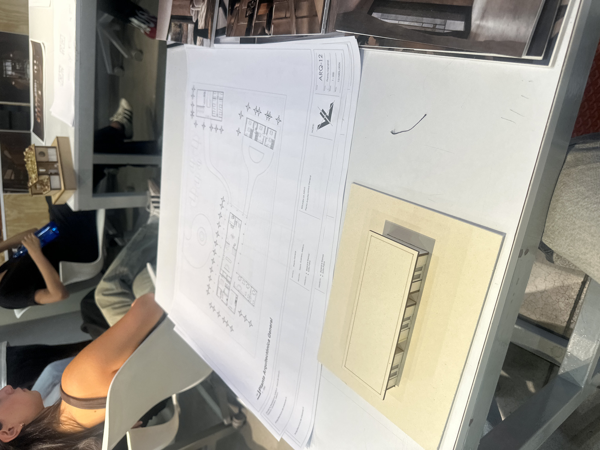The image shows a table or desk covered in architectural plans, models, and other papers. The plans are titled "Planta Arquitectónica General" (Architectural General Plan) and "Planta General" (General Plan), and include the date "15/ABRIL/2026". One of the plans has the identifier "ARQ-12". A simple model of a building is visible on the table. In the background, people are seated at tables. The location is identified as Mexico City, Mexico.