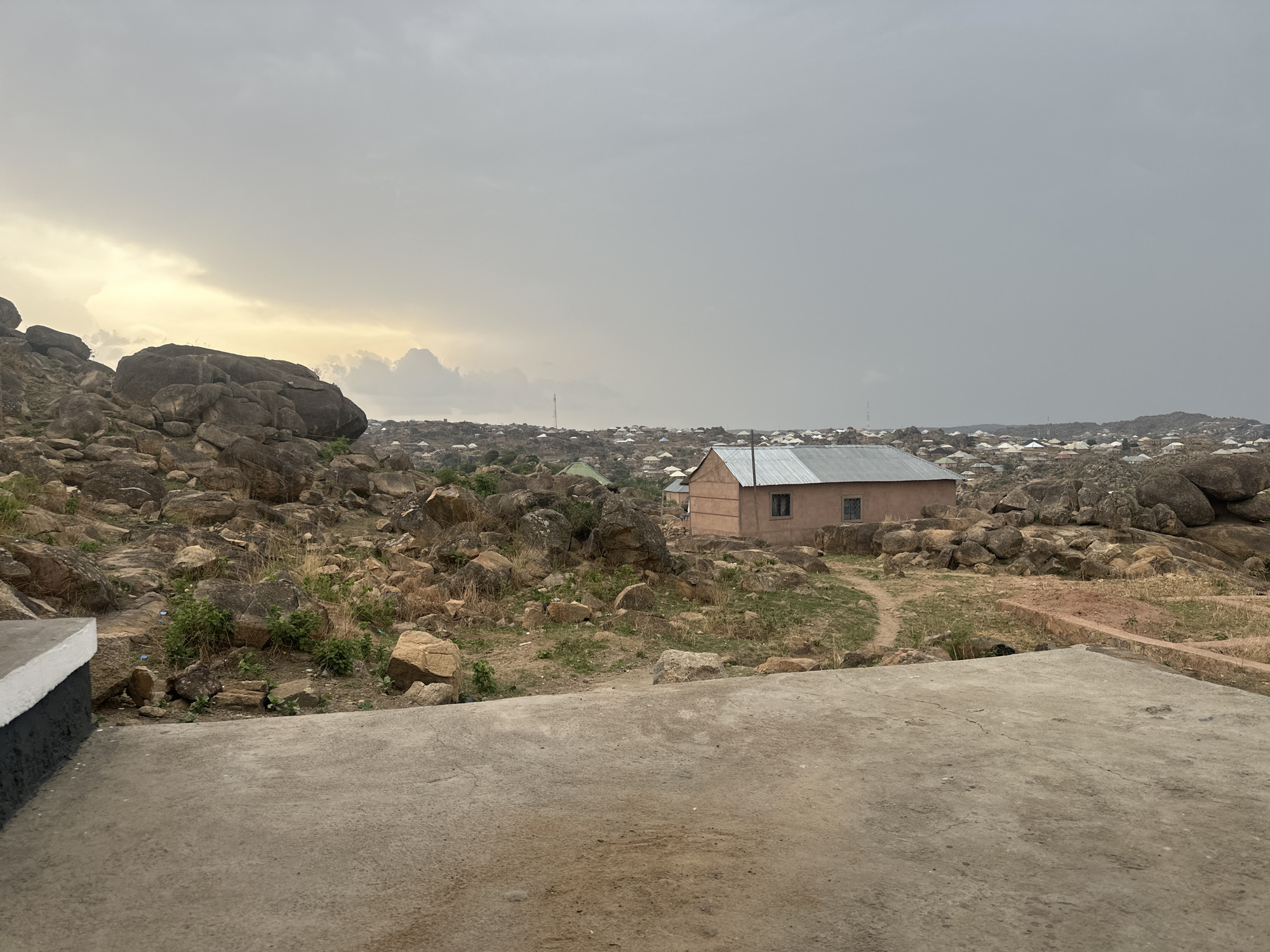 The image depicts a rocky landscape in Jos, Nigeria, under an overcast sky. In the midground, a peach-colored rectangular building with a corrugated metal roof stands. The surrounding terrain is covered with large boulders and sparse vegetation. In the background, a cluster of buildings with white roofs suggests a residential area. In the foreground, there is a concrete surface, possibly a patio or viewing platform, with a low concrete wall on the left edge, painted gray and white. The overall scene suggests a rural or peri-urban setting with a mix of natural and built environments.