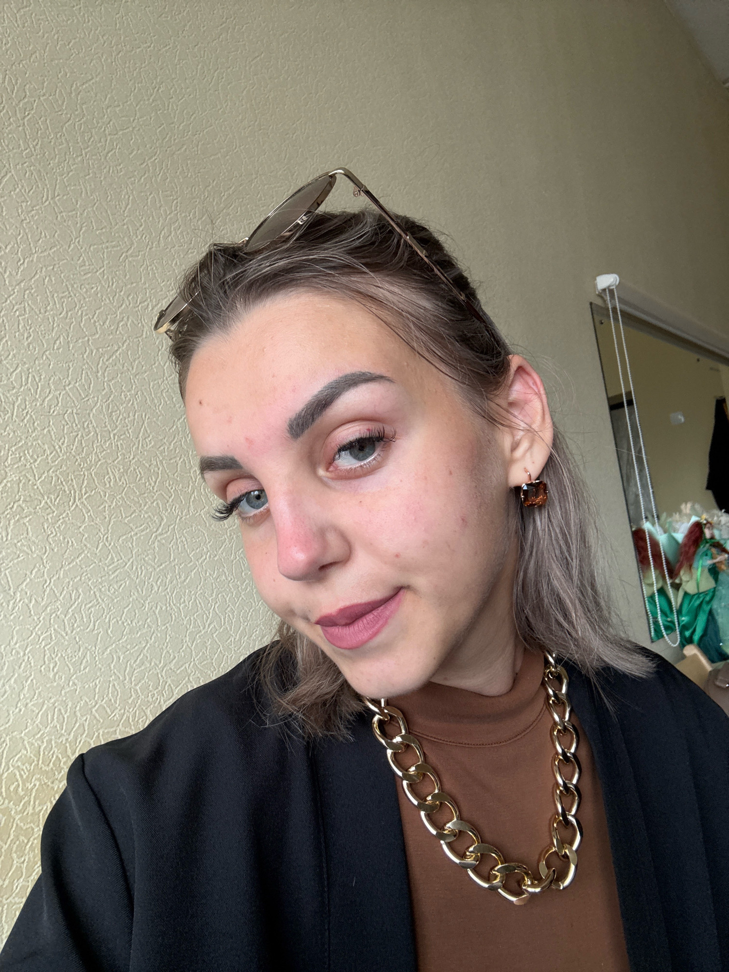 A young woman with fair skin, blue eyes, and shoulder-length blonde hair is shown in a close-up, selfie-style portrait. She is wearing a black blazer over a brown turtleneck and a chunky gold chain necklace. She also has a pair of sunglasses resting on her head and large, square, amber-colored earrings. Her makeup includes mascara, eyeliner, and a matte pink lipstick. The background appears to be an interior setting, with a textured beige wall on the left and a mirror reflecting items in the background on the right. The overall impression is a casual, fashion-conscious pose. There is no indication of the location in Yuzhno-Sakhalinsk, Russia.