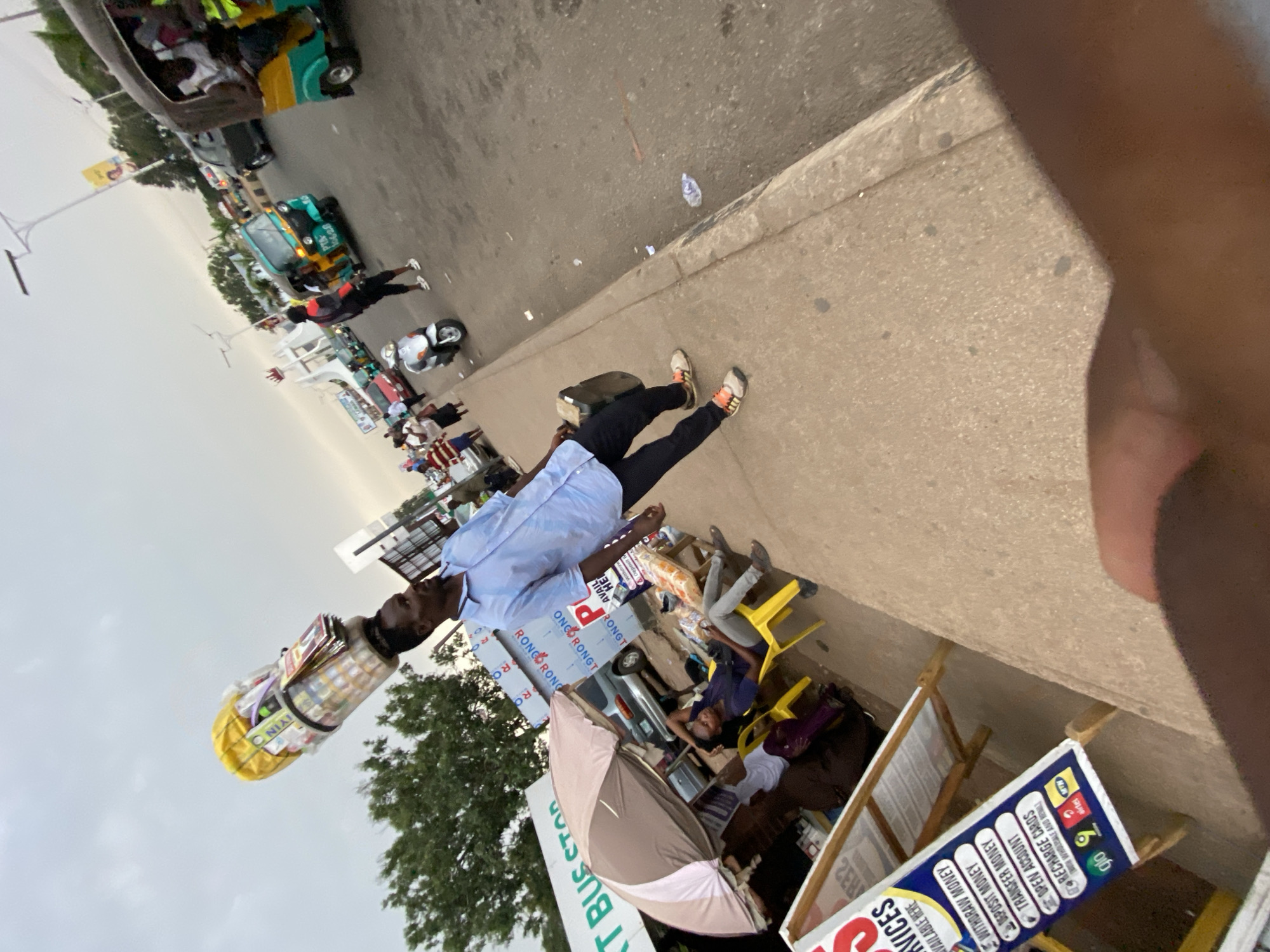 The image captures a bustling street scene, likely in Jos, Nigeria, given the signage and general environment. In the foreground is a sidewalk where some people are seated. Various posters advertise financial services, including money withdrawal, deposits, transfers, account opening, and recharge cards for mobile networks such as Glo, MTN, and Airtel.
A vendor with a container of goods on his head stands on the street. Several auto rickshaws, locally known as 'keke napep,' are parked, suggesting a transport hub or a busy commercial area. Other vehicles are also visible. The overcast sky suggests it could be morning or late afternoon.