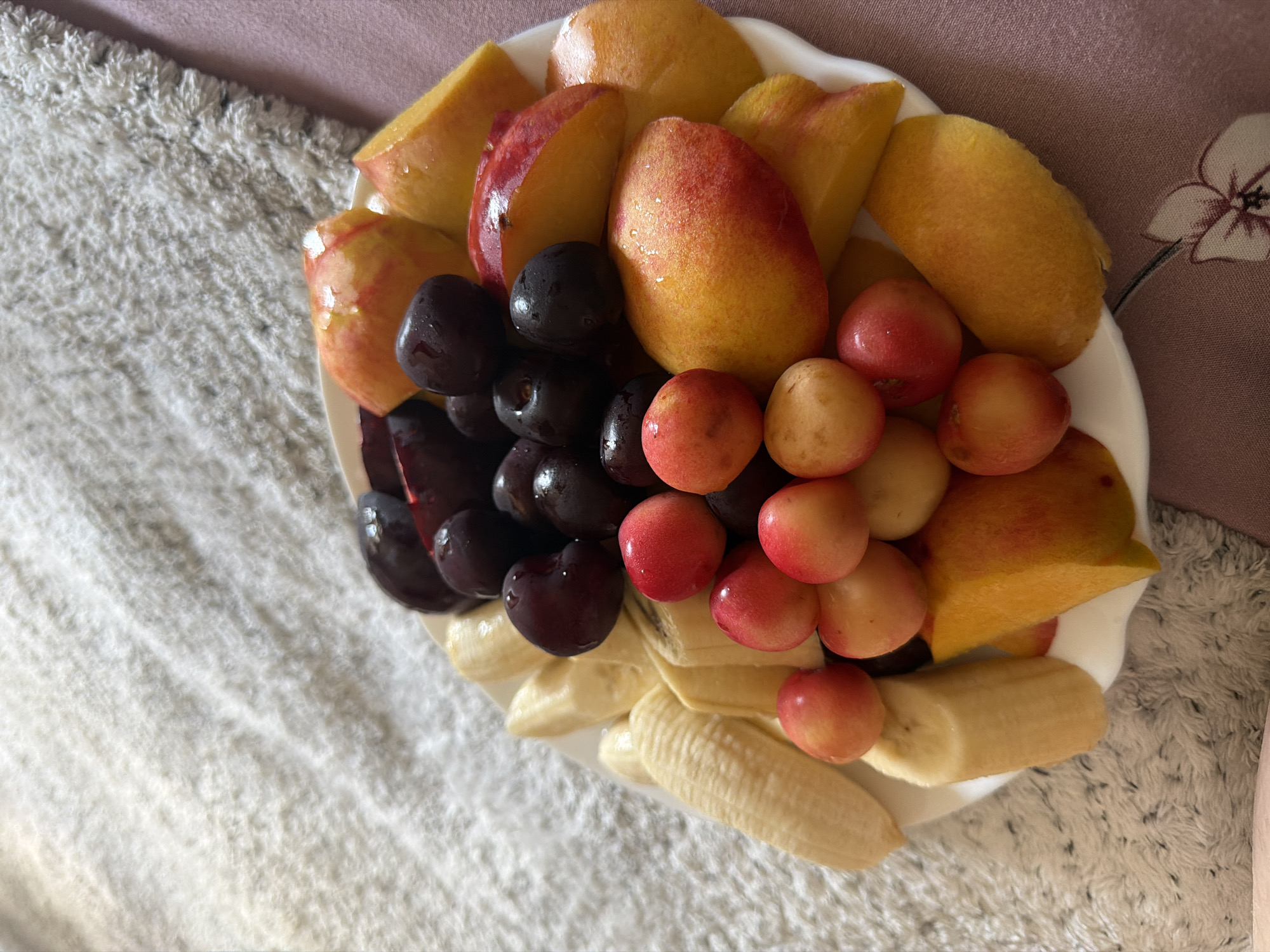 A close-up, overhead shot shows a white plate filled with a variety of cut and whole fruits, arranged on a textured, cream-colored surface. The fruits include sliced bananas, whole dark cherries, halved and quartered peaches or nectarines, and small, round red cherries. The fruits appear fresh and slightly dewy. The background consists of a soft, shaggy textile in a light color and a darker pink fabric with a floral pattern. There are no people or animals visible in the image. The lighting is soft and natural, casting gentle shadows. The image does not provide any information about the location in the city of Tbilisi, Georgia.