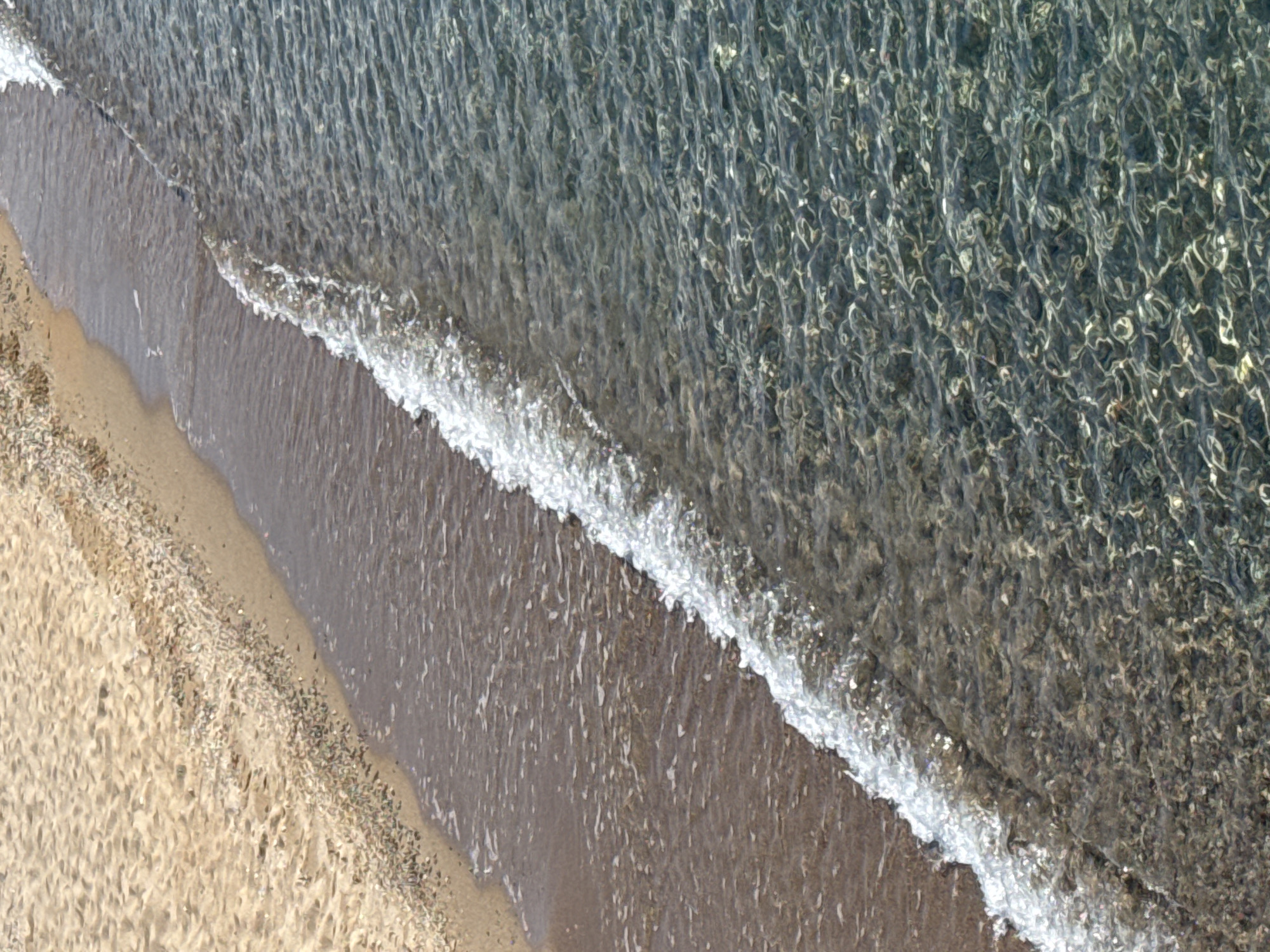 This is an aerial view of a sandy beach meeting clear, shallow water. The sand is light brown and textured with small pebbles and debris. A gentle wave is breaking on the shore, creating a line of white foam. The water is transparent, revealing the sandy bottom and the patterns created by the sunlight reflecting off the ripples. The scene is serene and natural, evoking a sense of tranquility. There is no visible text. The time of day appears to be sunny, as indicated by the bright reflections on the water. The location is Koshkol', Kyrgyzstan.
