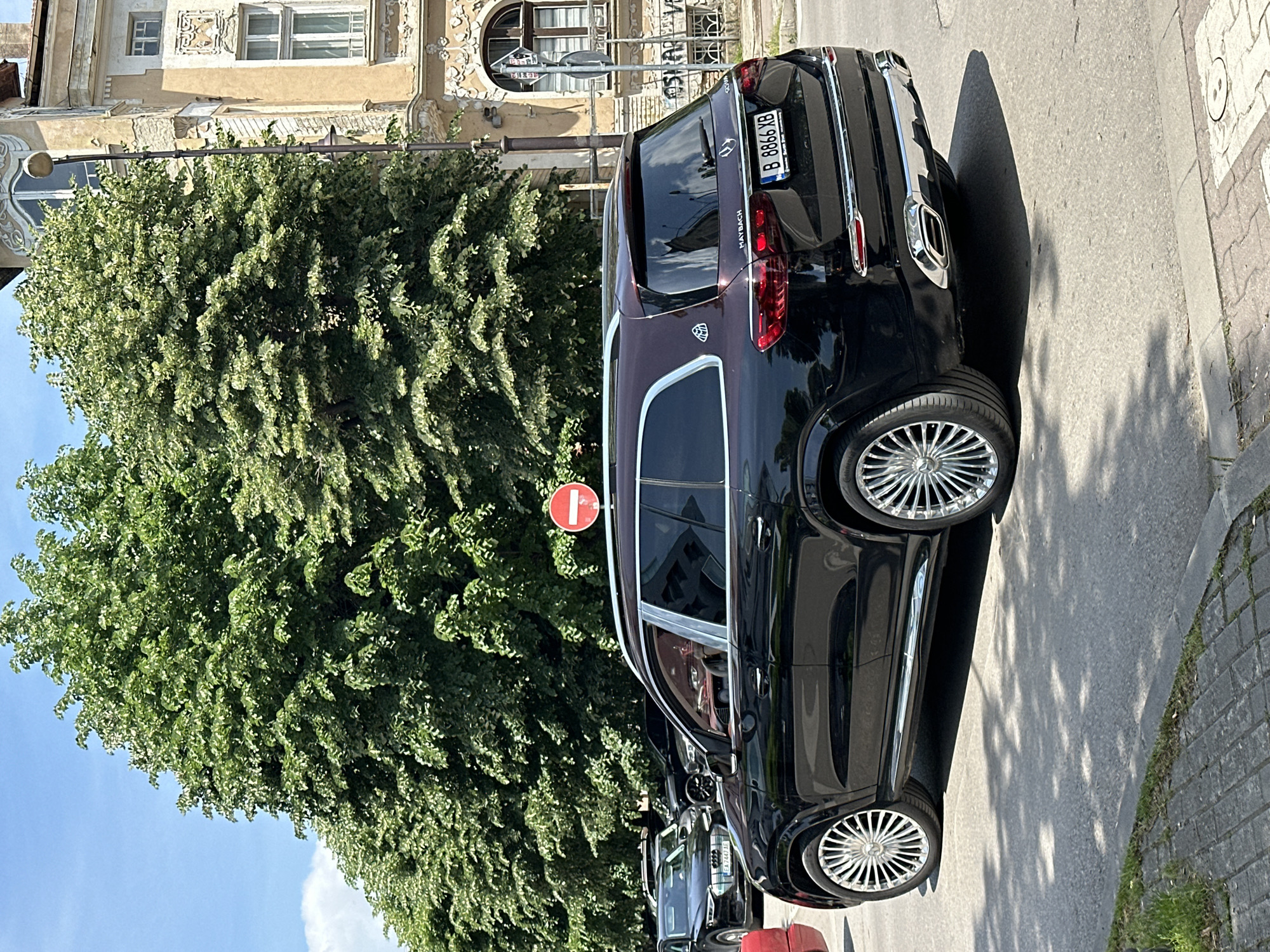 A black and burgundy two-tone Maybach SUV with chrome accents is parked on the side of a street in Varna, Bulgaria. The license plate number is B 8866 YB. A circular red and white no-entry traffic sign is positioned near the vehicle. A large, green tree occupies much of the left side of the frame. The building behind the tree features ornate architecture. The street is paved and the curb is visible on the right. Several other cars are partially visible behind the Maybach.