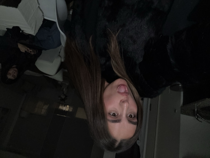A person with long brown hair and dark eyes sticks their tongue out at the camera. They are wearing a black turtleneck and black fur coat. In the blurry background, another person sits in a white chair next to a white bed. The room is dimly lit, and a window is visible in the distance.
