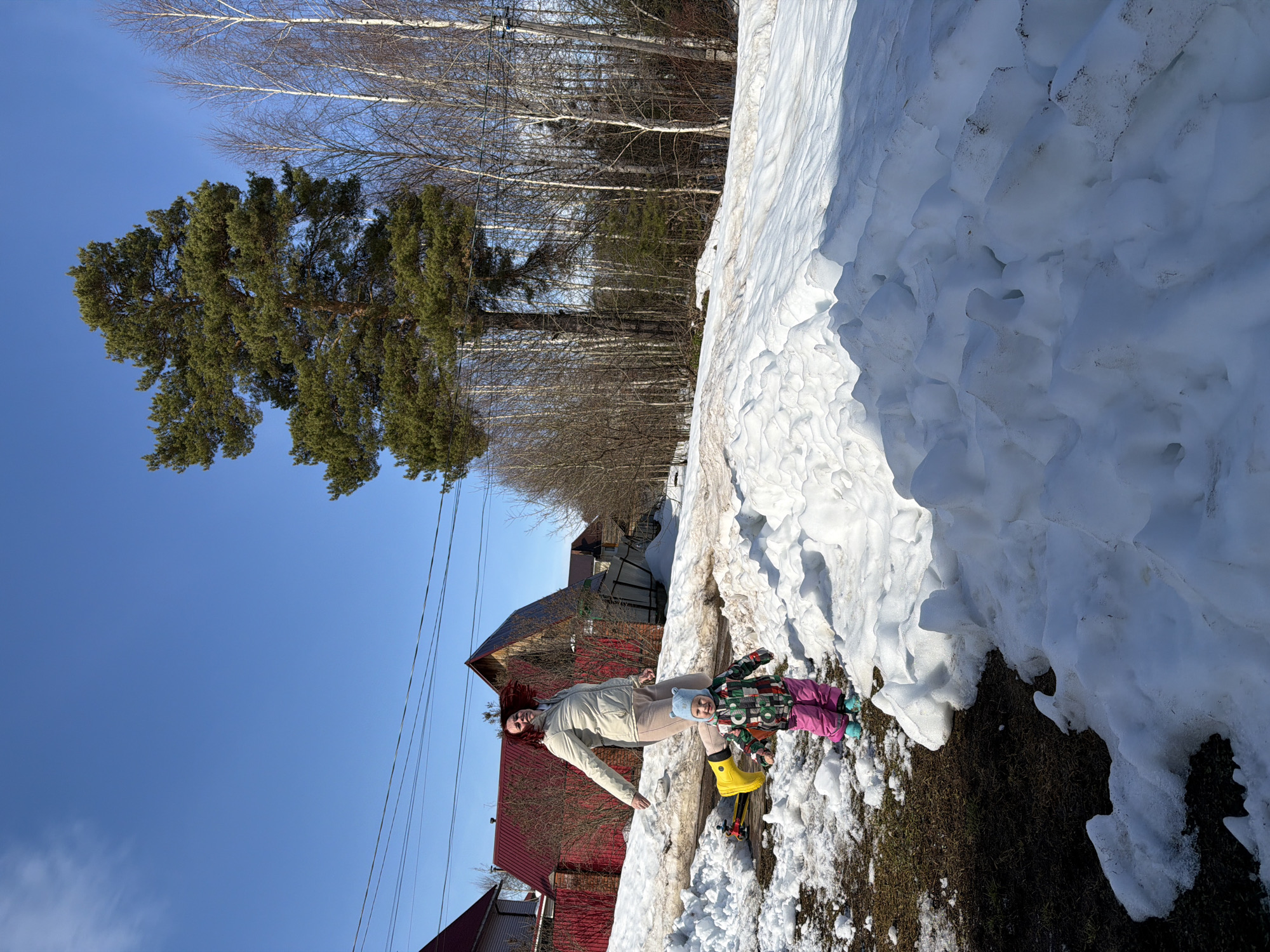 The image depicts an outdoor scene in Chaikovsky, Russia, characterized by significant snowmelt under a clear, bright blue sky. Two individuals are present: an adult female and a young child.
The adult female, with reddish-brown hair in motion, is captured mid-stride, appearing to jump or step over a low snowdrift in the middle ground. She wears a light-colored puffer jacket, light-colored pants, and bright yellow boots. Her gaze is directed towards the child.
The child, a toddler, stands in the foreground on a patch of dark earth adjacent to a snowdrift. The child wears a multi-patterned winter jacket, bright pink pants, light blue boots, and a light blue hat with ear-like features, and is smiling towards the camera. A yellow plastic shovel with a red handle is partially visible on the ground behind the child.
The ground consists of large, textured snowdrifts interspersed with patches of dark, exposed earth and sparse brown vegetation. In the left background, a house with a red roof and a red fence is visible. Overhead, utility wires traverse the sky. The right side of the background features a prominent, tall coniferous tree with dark green foliage, alongside numerous bare deciduous trees, primarily birches, forming a dense line suggestive of a forest edge. Sunlight is strong, casting visible shadows.