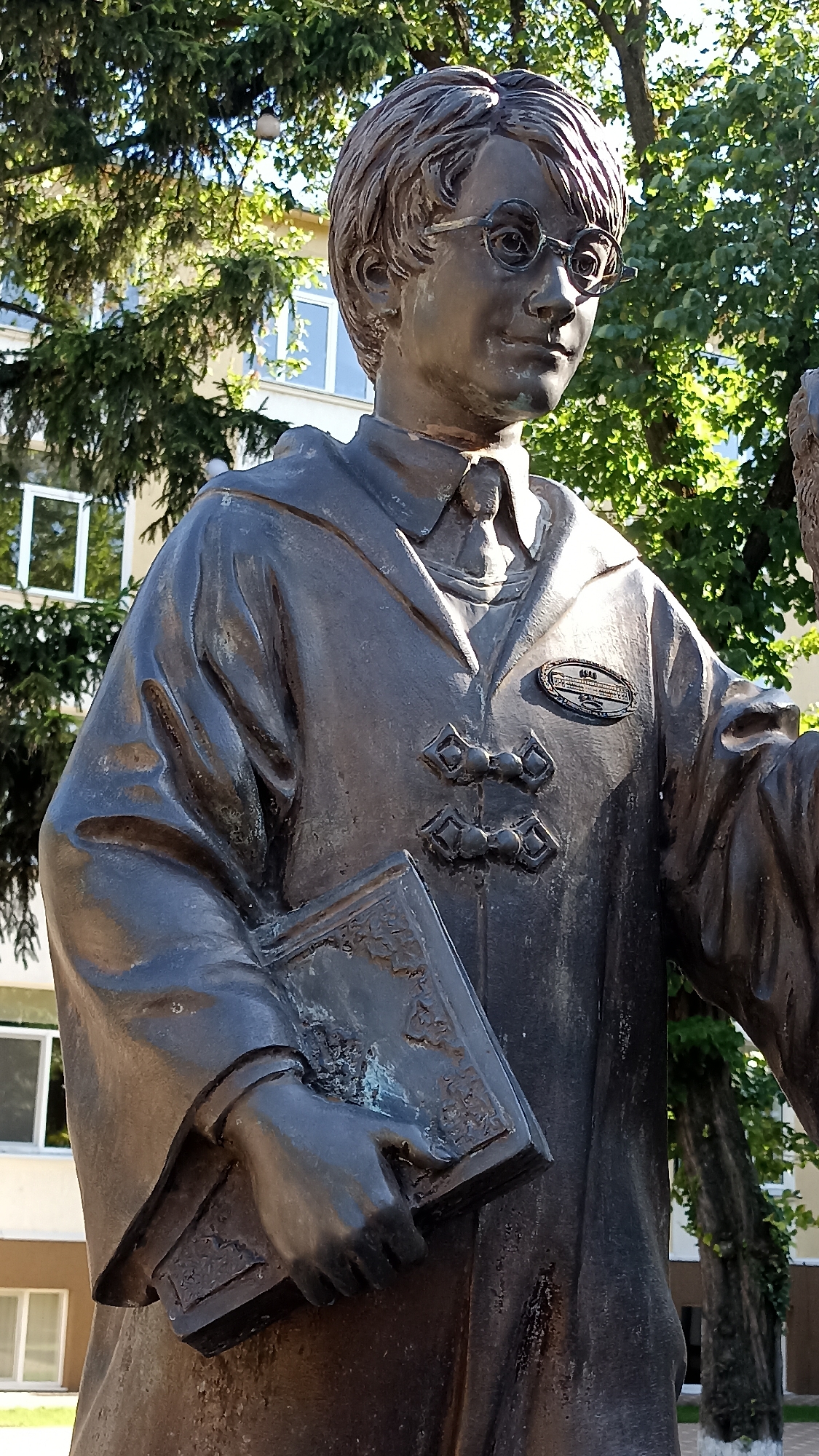 The image shows a bronze statue of a young boy with glasses, wearing a robe and holding a book. The statue is located in Tiraspol, Moldova, specifically in front of the Taras Shevchenko State University. The boy's expression is thoughtful, and his attire suggests he is a student. The statue is placed outdoors, with trees and buildings visible in the background. The lighting indicates it is daytime, with sunlight casting shadows. The overall impression is one of reverence for scholarship and youth.
