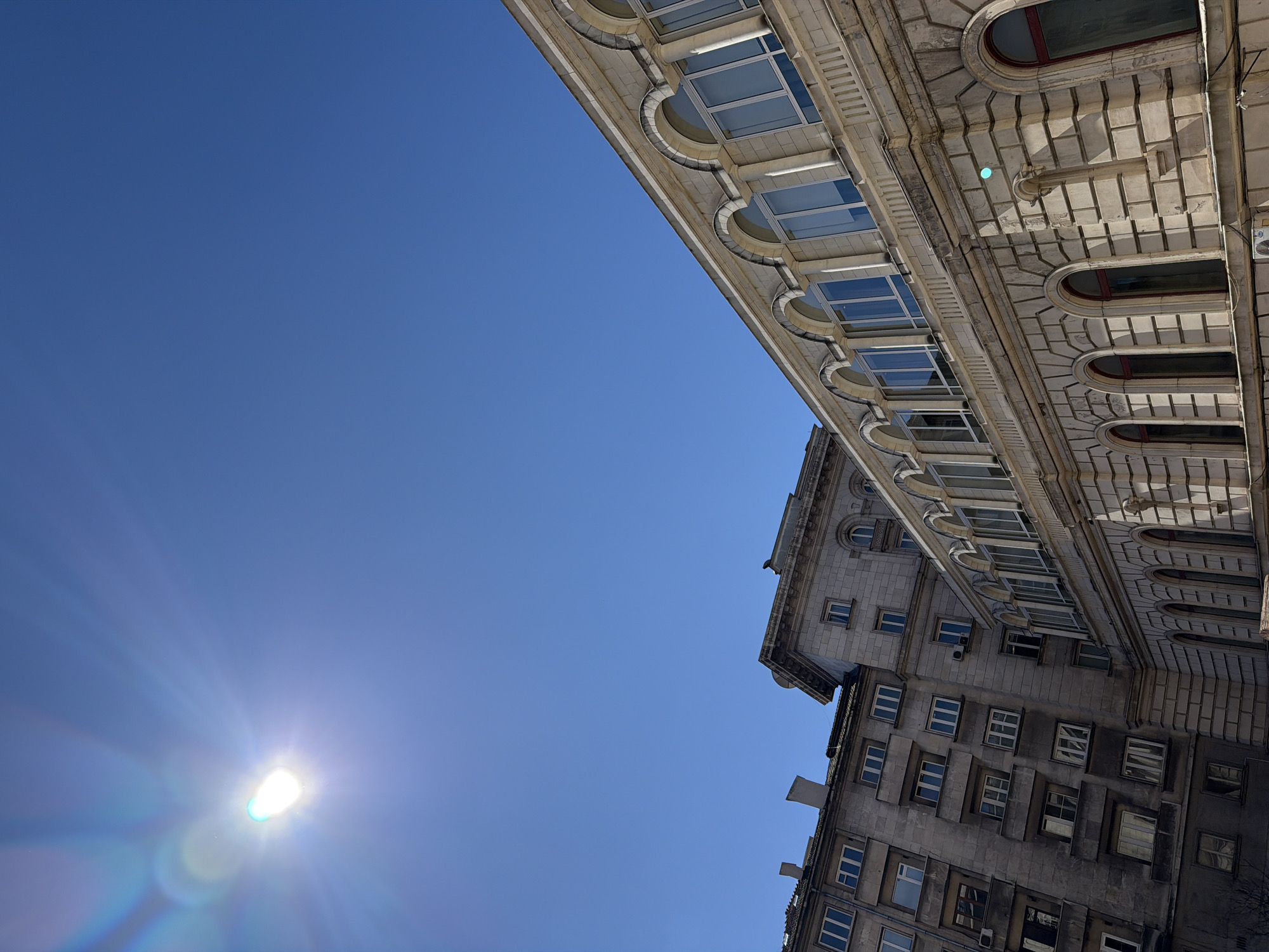 The image shows a low-angle view of buildings against a clear blue sky with a bright sun. The buildings feature architectural details like arched windows, columns, and decorative elements. One building has a lighter façade and the other has a darker, older look. The sun is visible in the lower left, causing a glare. The location appears to be an urban setting in Sofia, Bulgaria, judging by architectural styles.