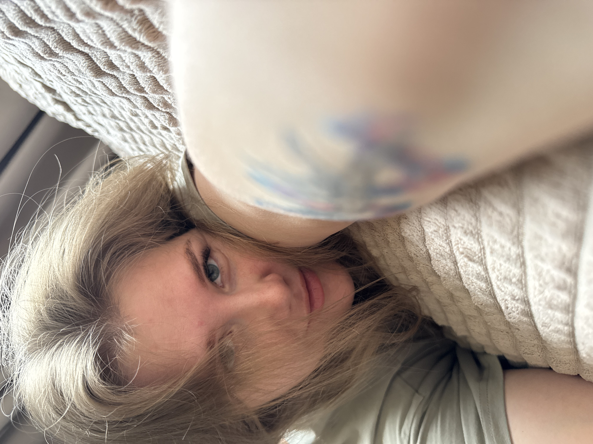 A low-angle, close-up photograph shows a woman with light blonde hair and blue eyes. She is lying down, with her head tilted towards the camera. A portion of her body, possibly an arm or shoulder, is in the foreground, partially obscuring the view. There is a tattoo visible on this body part. The background includes a textured, light-colored fabric, possibly a blanket or cushion. The location is not discernible from the image. No specific details relate to Gornyi (Dvurechenskoe S/P), Russia.
