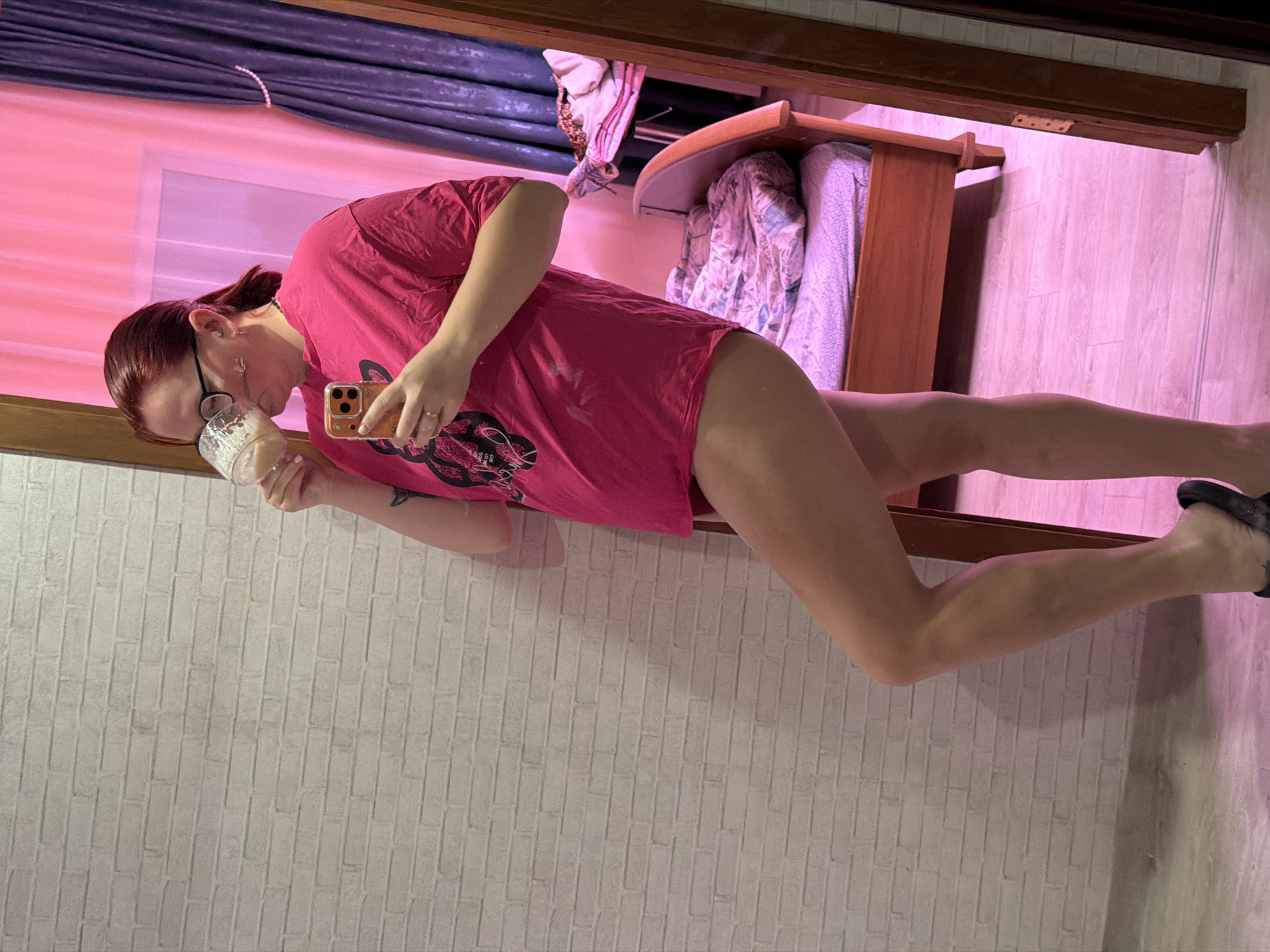 The image portrays a female individual in a domestic interior within Chaikovsky, Russia, captured in a full-length mirror reflection. The subject, with reddish-brown hair tied back, wears black-framed eyeglasses and features facial piercings near her mouth. A tattoo is visible on her upper left arm. She is dressed in an oversized, fuchsia-pink t-shirt with a black graphic, exposing her bare legs, and wears black slide sandals. Her right hand holds a clear glass mug containing a light-colored, foamy beverage, from which she is actively drinking. Her left hand holds an orange-cased smartphone, positioned to take the mirror selfie.
The background on the left features a white brick-patterned wall. To the right, the environment is bathed in a distinct pinkish-magenta light. This area reveals a window covered by a sheer pink curtain and a darker blue/purple curtain secured by a beaded tie-back. A wooden bed frame with unmade, patterned bedding is visible to the right of the subject, next to a surface holding folded garments. The flooring consists of light-colored wood or laminate.
