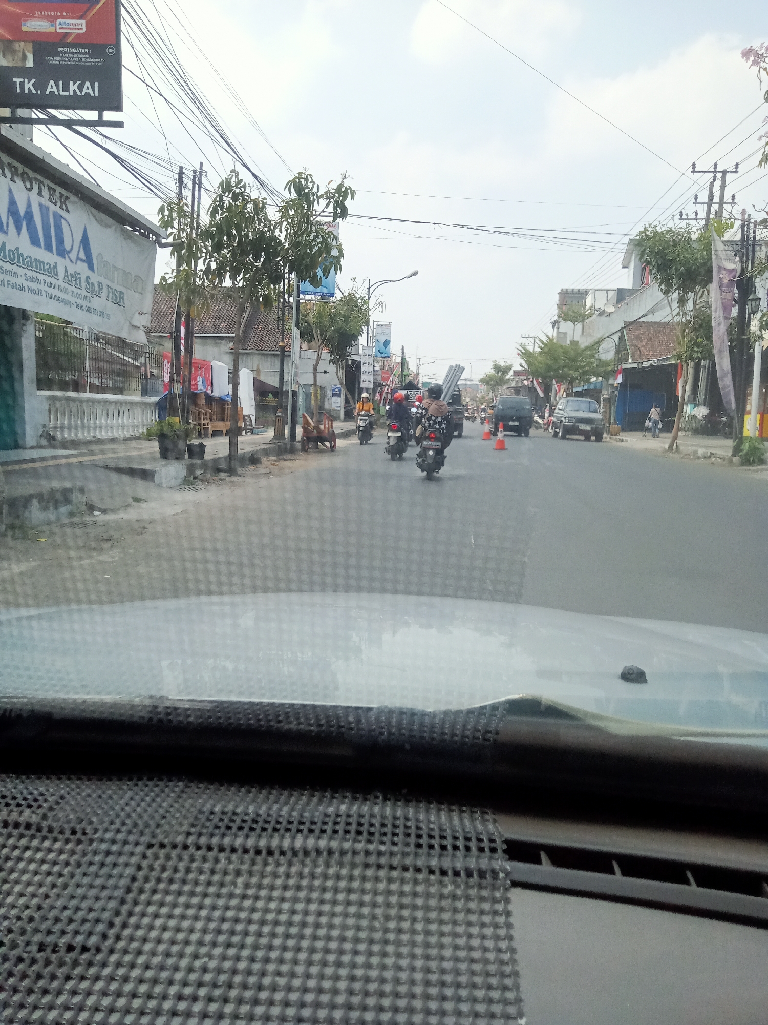 The image captures a street scene in Mangunsari, Indonesia, viewed from inside a vehicle. The street is lined with buildings and trees, with a clear sky overhead. Multiple vehicles, including motorcycles and cars, are present on the road, some in motion. People are visible riding motorcycles and walking. A sign for "TK. ALKAI" is prominently displayed on the left side of the street, along with another sign that appears to be for a pharmacy. Electrical wires crisscross above the street. The foreground shows the interior of the vehicle, including the dashboard and windshield. The scene suggests a typical day in a busy urban environment in Indonesia.