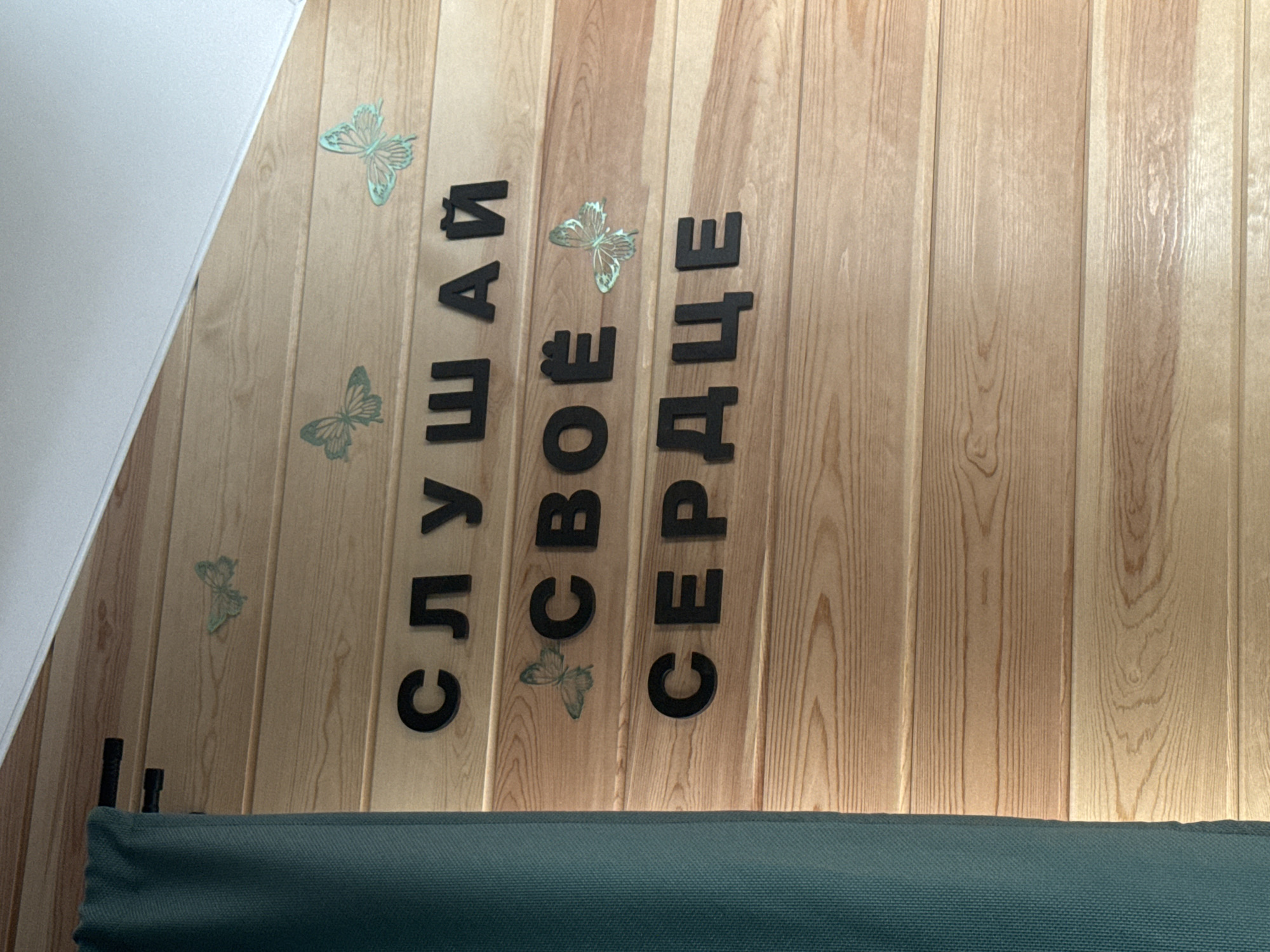 The image shows a wooden wall with black lettering spelling out "СЛУШАЙ СВОЁ СЕРДЦЕ" which translates to "LISTEN TO YOUR HEART" in Russian. There are several decorative butterflies placed on the wall above and between the words. The wall is made of horizontal wooden planks, and a teal curtain is visible on the left side of the image, obscuring part of the wall and the full extent of the lettering. The scene appears to be indoors, possibly in a room or establishment in Khuzhir, Russia.