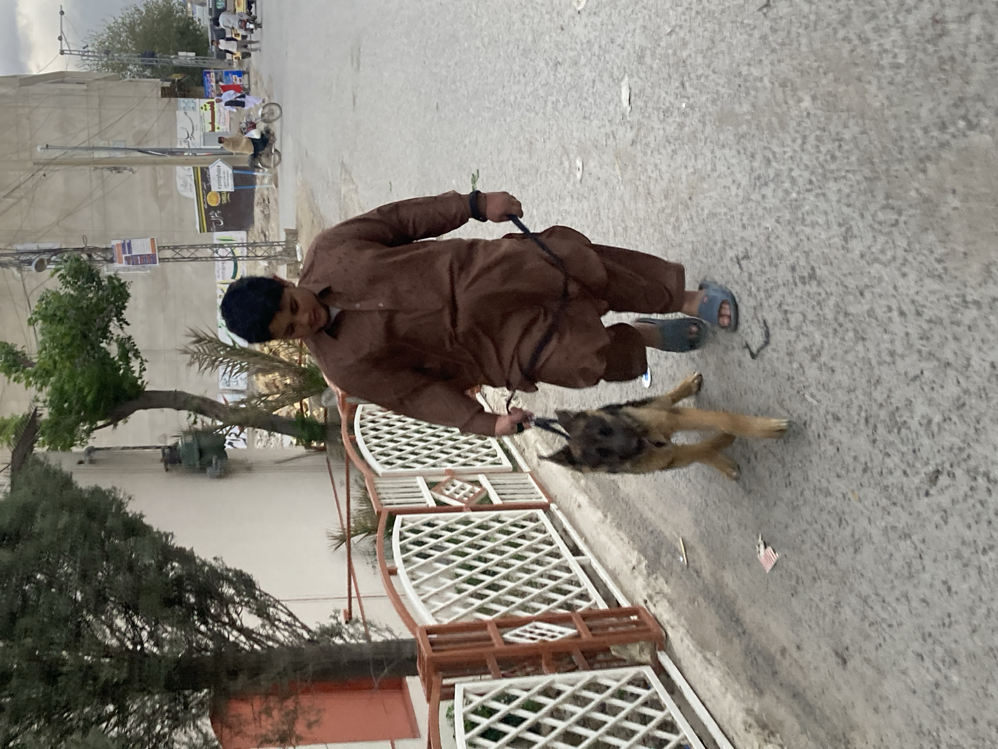 A man wearing a brown shalwar kameez and blue sandals walks a dog on a leash in Quetta City Tehsil, Pakistan. The man is walking on a paved road. A white and red fence is visible to the left, along with a tree. In the background, there are buildings, trees, and people on motorcycles. The sky is overcast.