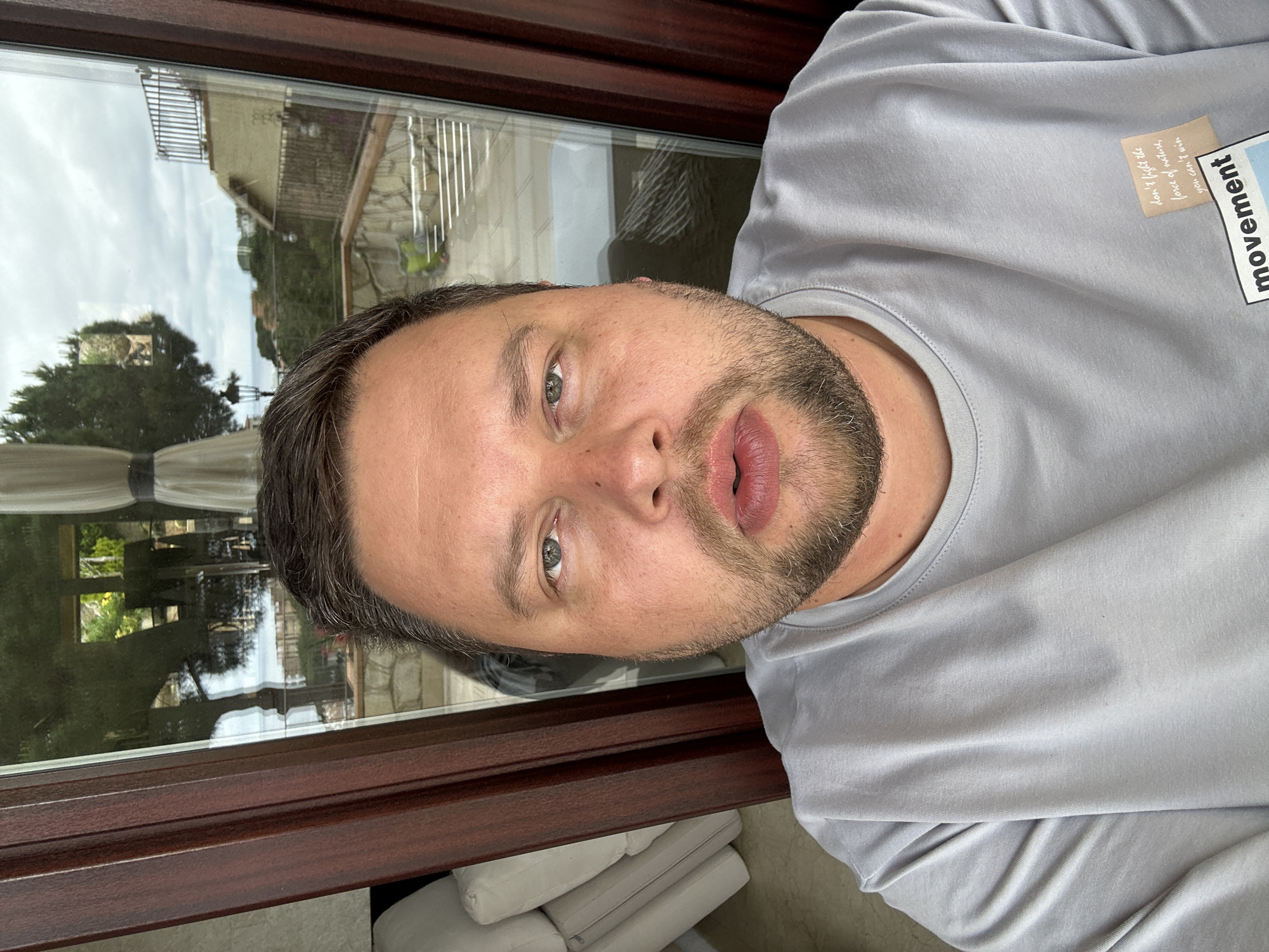 A man is positioned indoors, likely near a window. The location is plausibly Sant Feliu de Guíxols, Spain based on the reflection in the window of a coastal landscape with Mediterranean architecture. The man has short brown hair, a stubble beard, and is wearing a gray T-shirt with a patch that reads "movement". The image is slightly rotated. There are pillows visible near the window frame.