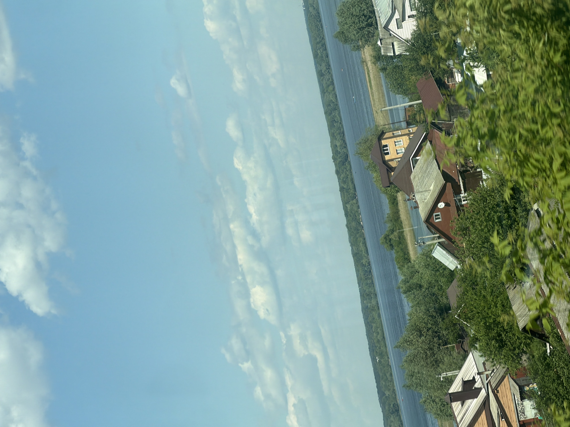 The image depicts a village with houses nestled among trees, overlooking a wide river under a partly cloudy sky. The architecture suggests a rural Russian setting. Specifically, this view is characteristic of the village of Nizhniye Vyazovye, Russia, a location known for its proximity to the Volga River, which is visible in the background. The houses appear to be a mix of older, traditional structures and more modern dwellings, with various roof styles and materials evident. Lush green trees and vegetation surround the houses, and the river reflects the blue sky and clouds. There are no people visible in the image, nor are there any apparent actions or interactions taking place. The scene is peaceful and tranquil, conveying a sense of everyday life in a riverside community.