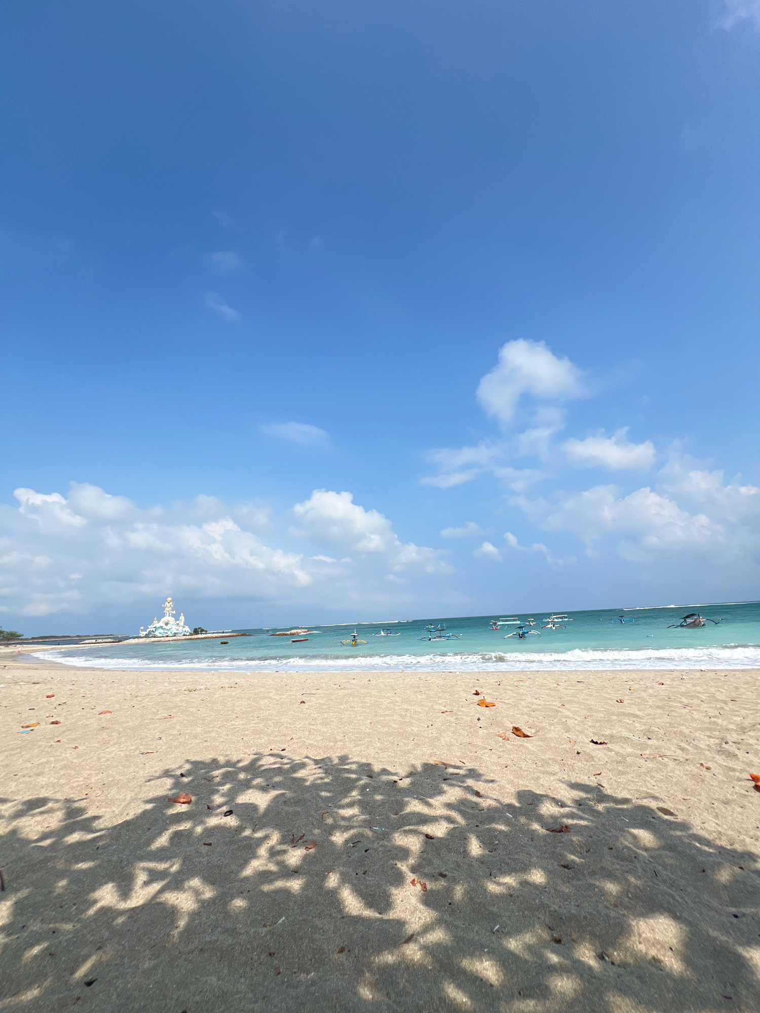 This is a ground-level, wide-angle shot of Kuta Beach in Bali, Indonesia. The foreground is dominated by a sandy beach with scattered dry leaves and shadows of foliage cast from the upper right. The middle ground features the ocean with gentle waves lapping at the shore. Several traditional Balinese fishing boats, known as jukungs, are visible on the turquoise water. In the distance, to the left of the frame, is a large white monument, possibly a statue or temple structure, situated on a rocky outcrop near the coastline. The sky above is a clear, bright blue with scattered white cumulus clouds. The scene appears serene and tranquil, with no people clearly visible on the beach or in the water.
