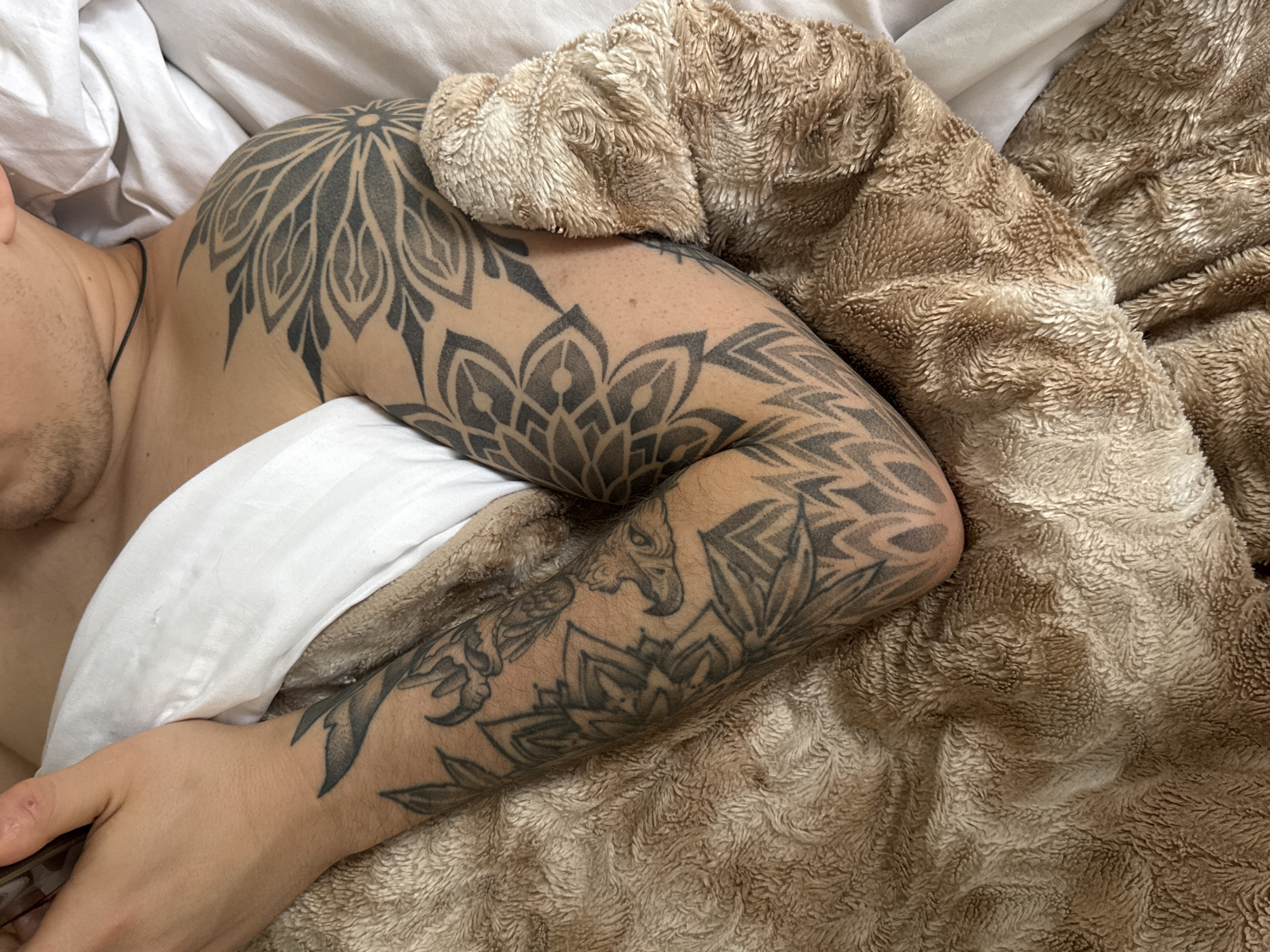 An overhead shot shows a person, likely male, lying in a bed, partially covered by white bedding and a fluffy brown blanket. The person's left arm and shoulder are extensively covered in black and grey tattoos.
The tattoos feature intricate designs, including a prominent large, circular mandala-like pattern on the shoulder, and various geometric and stylized floral or tribal patterns extending down the length of the arm. Some patterns on the lower arm suggest elements resembling mythical creatures or animals integrated into the overall design.
The individual is shirtless, with a thin black cord necklace visible around their neck. Their face is mostly out of frame, but a portion of their chin and neck is visible, suggesting a relaxed posture, possibly resting or sleeping. The white bedding appears soft and wrinkled, while the brown blanket has a deeply textured, plush, and cozy appearance. The scene is illuminated by soft, ambient light, implying an indoor setting. There is no visible text in the image. This scene takes place in Rostov-on-Don, Russia.