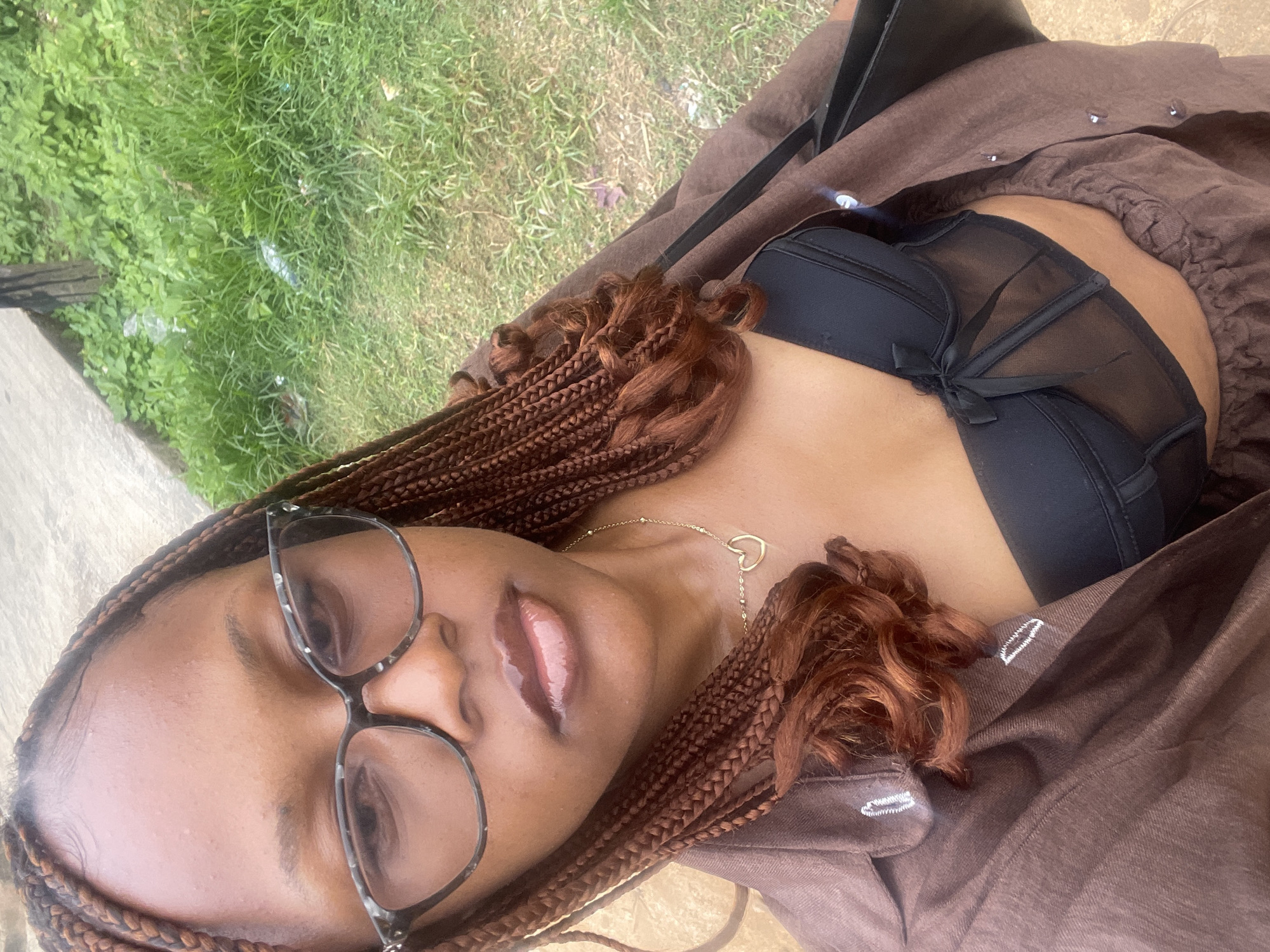 A young woman with braided hair and glasses takes a selfie outdoors, with a grassy area and a paved surface visible in the background. She is wearing a brown open shirt over a black bustier and a brown skirt. A simple gold heart necklace is visible around her neck. The lighting suggests it is daytime. The location is identified as Rafin Par, Nigeria.