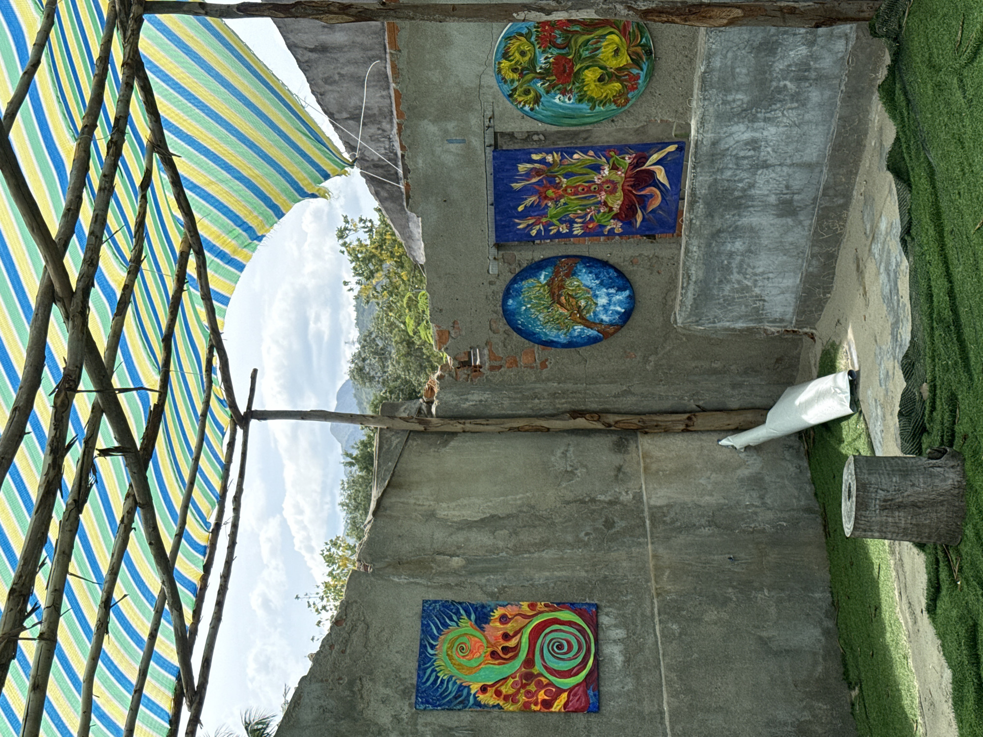 In Vĩnh Lương, Vietnam, an outdoor art display is set up under a striped awning. The awning, composed of alternating blue, green, and yellow fabric strips, is supported by wooden poles and branches. Against a weathered concrete wall, four distinct artworks are mounted. On the left, a rectangular painting features a swirling, vibrant green and orange abstract design. To its right, a circular painting depicts a stylized tree with a blue background. Next to it is a rectangular painting with a blue background showcasing a red and yellow floral arrangement. Finally, on the far right, another circular painting displays a cluster of yellow and red flowers against a teal background. The ground is covered in artificial grass, and a rough-hewn tree stump sits on the left. A white rolled-up sheet of material is leaning against the wall near the stump. The background reveals a natural landscape with trees and hills under a cloudy sky, with the rough, broken edges of the surrounding structure suggesting an unfinished or rustic construction.