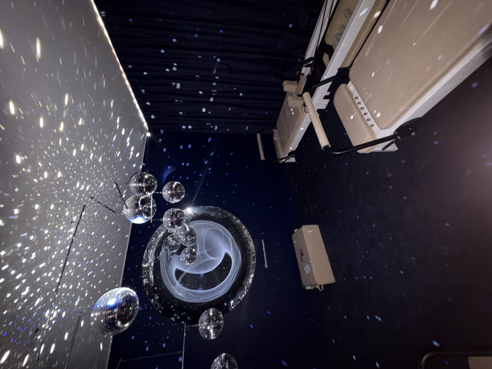 In a dimly lit room, possibly a studio or gym, several disco balls hang from the ceiling, casting sparkling reflections across the dark blue walls and floor. The walls are adorned with a large, circular projection of a swirling white graphic. Two Pilates reformers are positioned in the foreground, their light beige upholstery contrasting with the dark surroundings. A tan rectangular box sits on the floor to the left. The environment suggests a unique and possibly artistic fitness or event space. The location is identified as Mexico City, Mexico.