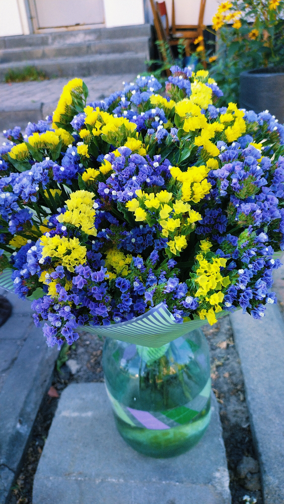 A bouquet of blue and yellow flowers is displayed in a green glass vase on a gray, concrete surface. The bouquet is wrapped in green and white striped paper. In the background, steps lead up to a building, and there are glimpses of foliage and a black planter. The scene appears to be outdoors, possibly in a public space or garden. The image is taken from a high angle, looking down at the flowers and vase. There are no people or animals visible in the image.