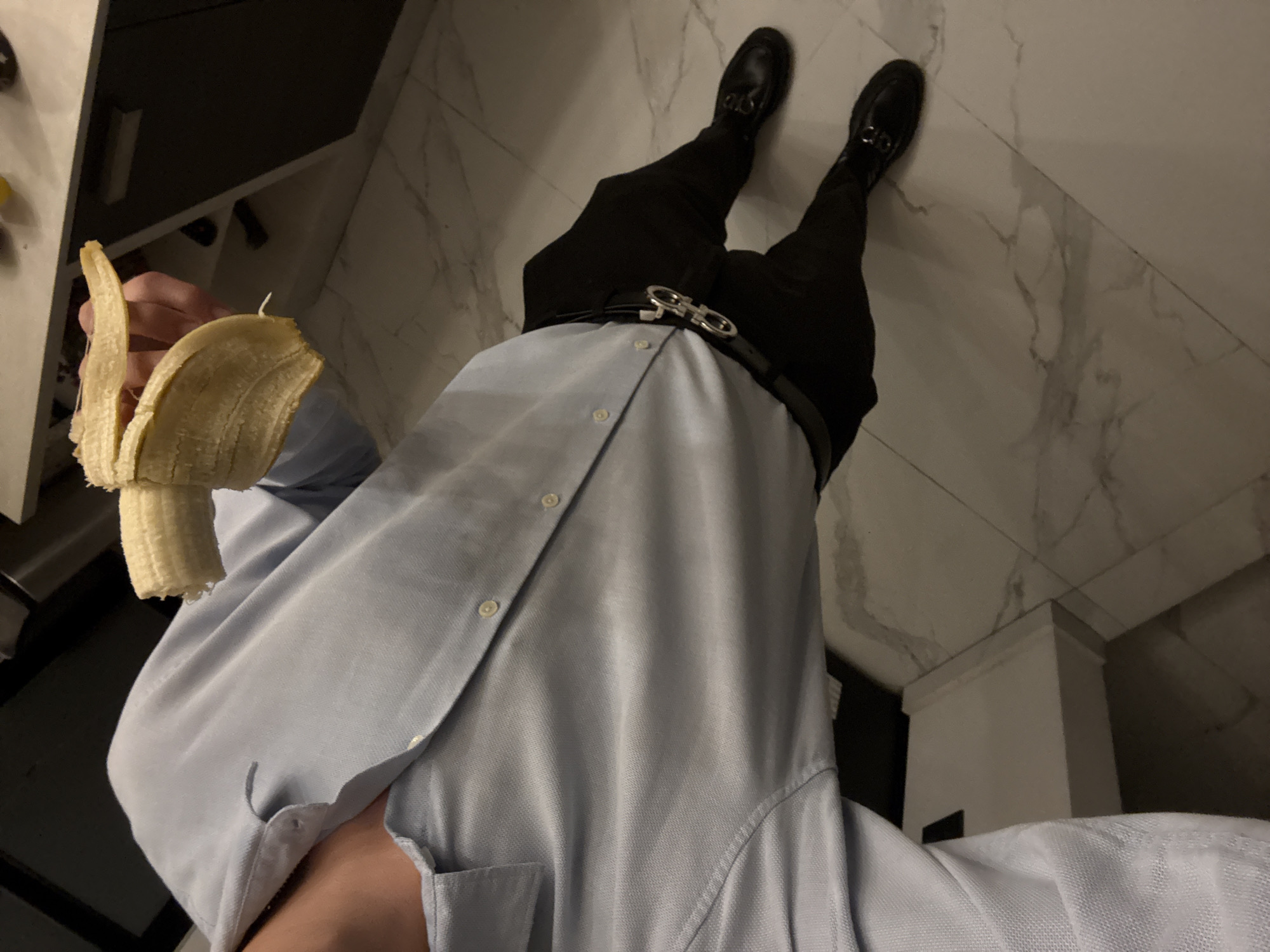 A low-angle shot from an indoor location with marble-like tiled flooring. A person is visible from the knees up, wearing black trousers, black shoes, and a silver belt buckle. A light blue button-down shirt covers the person's torso. The person holds a partially peeled banana in their left hand. The background includes a dark cabinet. The location is not in San Bernardino Tlaxcalancingo, Mexico.