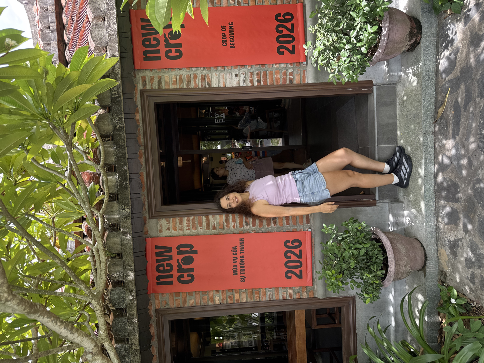 A woman stands to the right of a building entrance in Hoi An, Vietnam. She is leaning against a red banner that reads "new crop" in large black letters, with smaller text below, and "2026" at the bottom. Another similar banner is on the left side of the entrance. The building has a brick facade and a tiled roof. Two potted green plants flank the entrance. The woman is wearing a lavender tank top and denim shorts, with black athletic shoes. She is looking to her left and smiling. Sunlight casts shadows from trees and foliage onto the ground and building. Inside the building, through the open doorway, other patrons can be seen seated at tables.