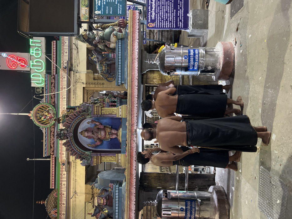The image captures a nighttime scene at a Hindu temple, likely in Palani, India, given the contextual information and visible text. Several bare-chested men, wearing black dhotis or lungis, stand with their backs to the viewer, appearing to be devotees engaged in prayer or waiting in a queue. Their posture suggests reverence as they face deeper into the temple complex.
The background features ornate and colorful temple architecture, including sculptures of deities such as Ganesha and Hanuman, adorning a gopuram-like structure. A bright neon sign spelling "முருகா" (Muruga) in Tamil is prominent on the left, indicating the primary deity. Below, other intricate carvings and smaller shrines are visible.
In the mid-ground, two large, cylindrical donation boxes (Hundis) are present. They are silver-colored with blue labels that read "பிரார்த்தனை உண்டியல்" (Prarthanai Hundi, meaning Prayer Donation Box) and "HUNDI". A small number '3' is also visible on one of the donation boxes.
Several informational signs in Tamil are affixed to pillars and walls, with text including "அருள்மிகு தண்டாயுத" (Arulmigu Dhandayuthapani, referring to the temple's presiding deity), "பக்தர்களுக்கு" (to devotees), and details about timings (e.g., "மாலை 4.00" and "மாலை 6.00 மணிக்கு") and devotee entry, suggesting operational instructions for visitors. The visible details, including the attire of the men, the temple architecture, and the Tamil script, strongly confirm a South Indian temple setting.