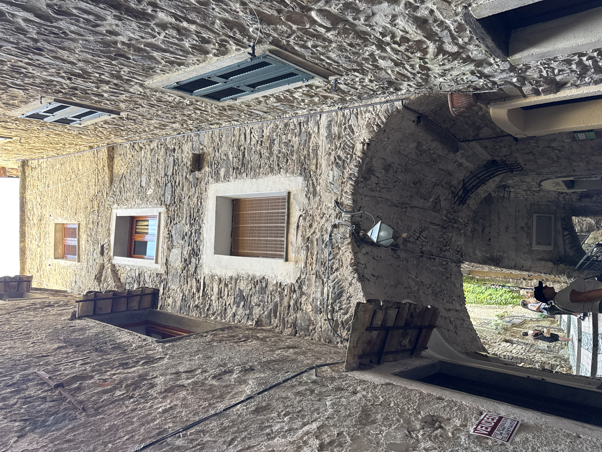 The image shows a narrow, stone-paved alleyway in Dolceacqua, Italy. The alley is lined with old stone buildings, some of which have wooden shutters on the windows. There is an arched passageway in the foreground with a light fixture hanging from the ceiling. In the background, a few people are walking up the alley, and there is a sign that says "VENDESI" (For Sale) on the left wall. The buildings are made of rough-hewn stones, and the overall impression is one of age and rustic charm. Sunlight filters down into the alley, creating areas of light and shadow.