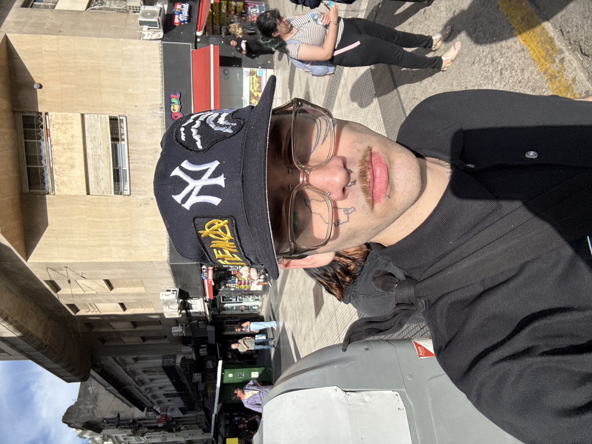 A close-up, slightly rotated, selfie-style photograph captures a person with piercings and tattoos. The person wears a black baseball cap with a New York Yankees logo and clear-framed glasses. The person is wearing a black shirt, and their hair is visible underneath. Behind the person, a street scene unfolds. A woman in a striped shirt and black pants stands near a building with shops. Other pedestrians are visible in the background. Buildings line the street, and the sky is visible.
The scene is identified as Buenos Aires, Argentina, by a sticker on a utility box.